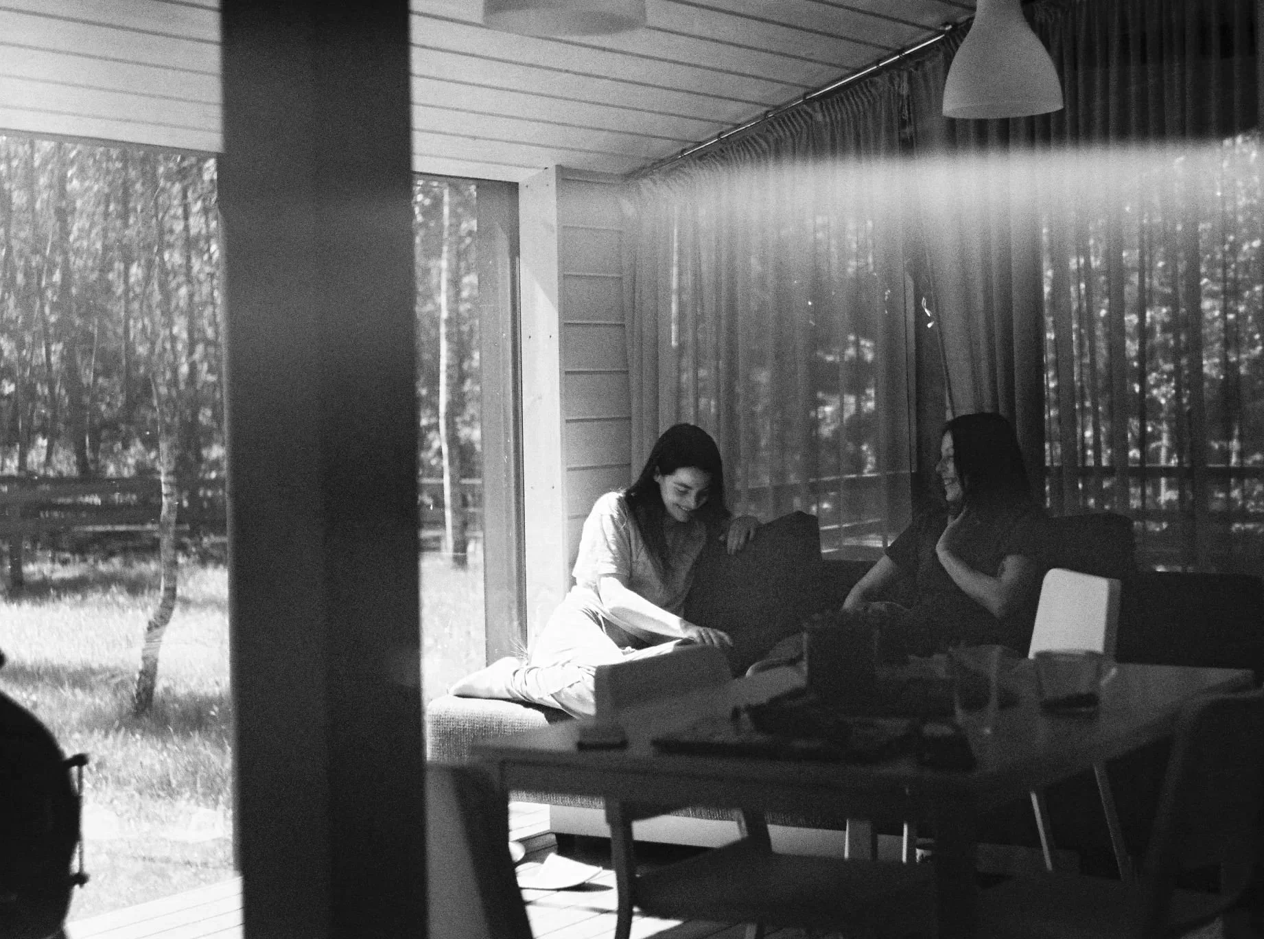 Two women sitting on a sofa indoors near large windows, engaging in conversation, with a forest visible outside.