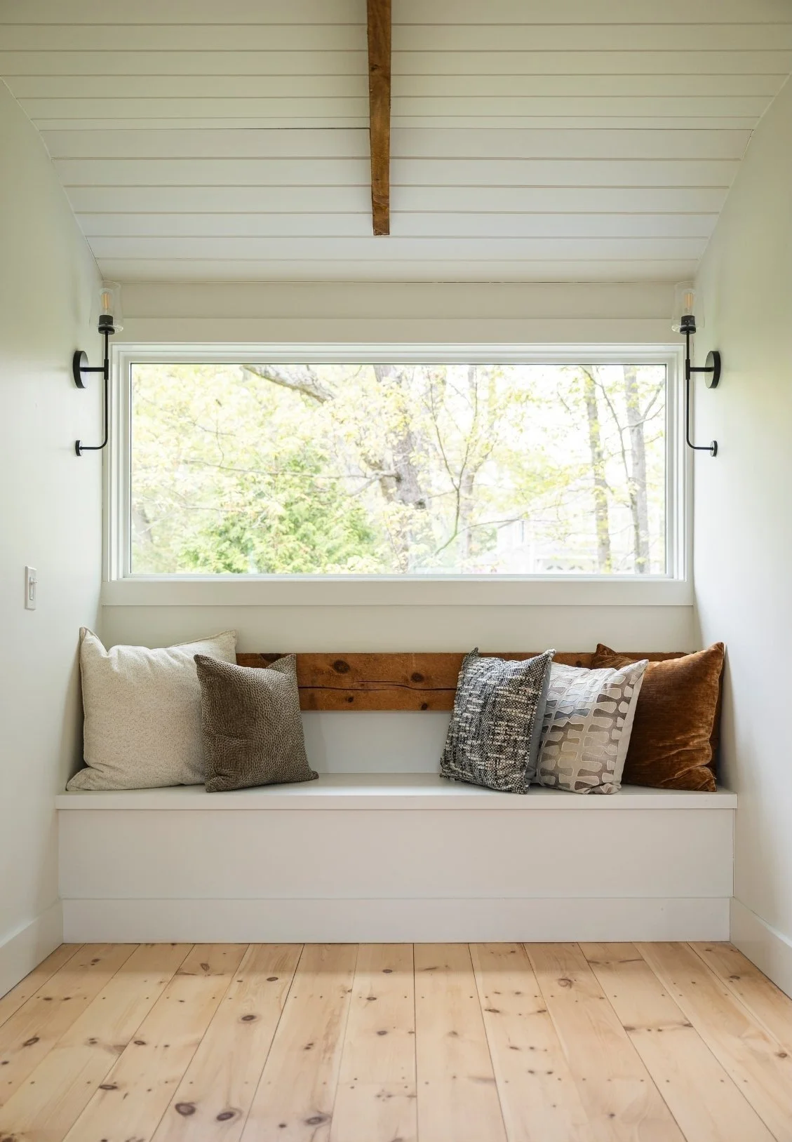 A cozy window seat with five decorative pillows against a large horizontal window, overlooking trees outside. Wall sconces are mounted on each side of the window, and the ceiling is white with wooden beams. The floor is light-colored wood.