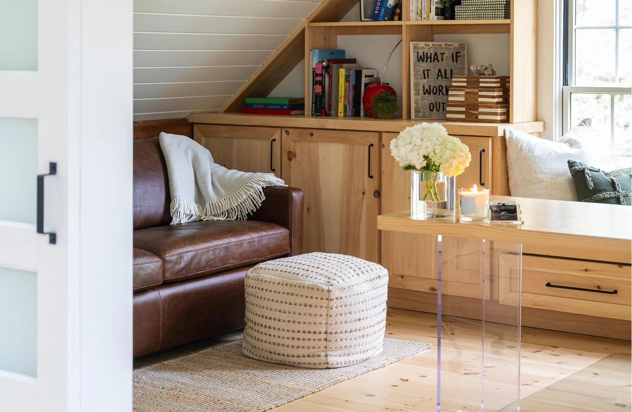A cozy living room with a brown leather sofa, a white fringed throw blanket, a striped round ottoman, and a wooden bookshelf filled with books and decor. There is a vase of white flowers on a glass table and a window with daylight streaming in.