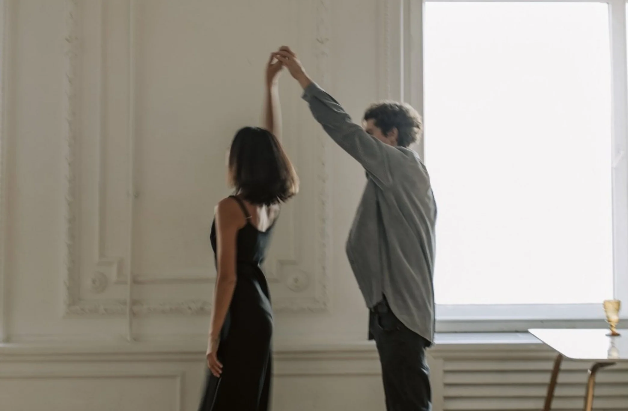 A man and a woman standing in a room with ornate white walls and large windows, facing each other and holding hands above their heads.