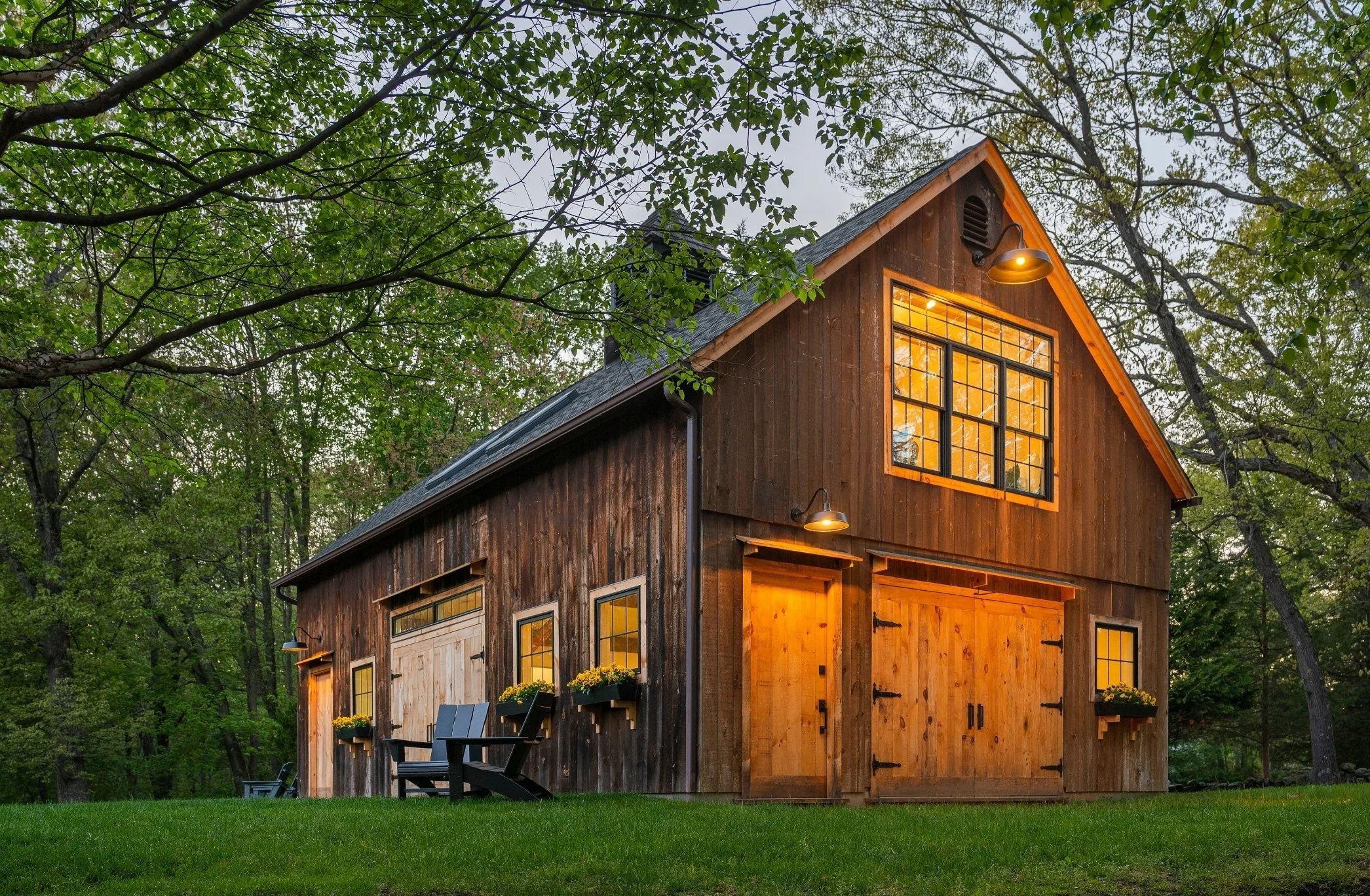 A wooden cabin surrounded by trees with a large window that has warm interior lighting. Outdoor patio furniture and potted plants are visible outside, and the scene is set during dusk or evening with an overcast sky.