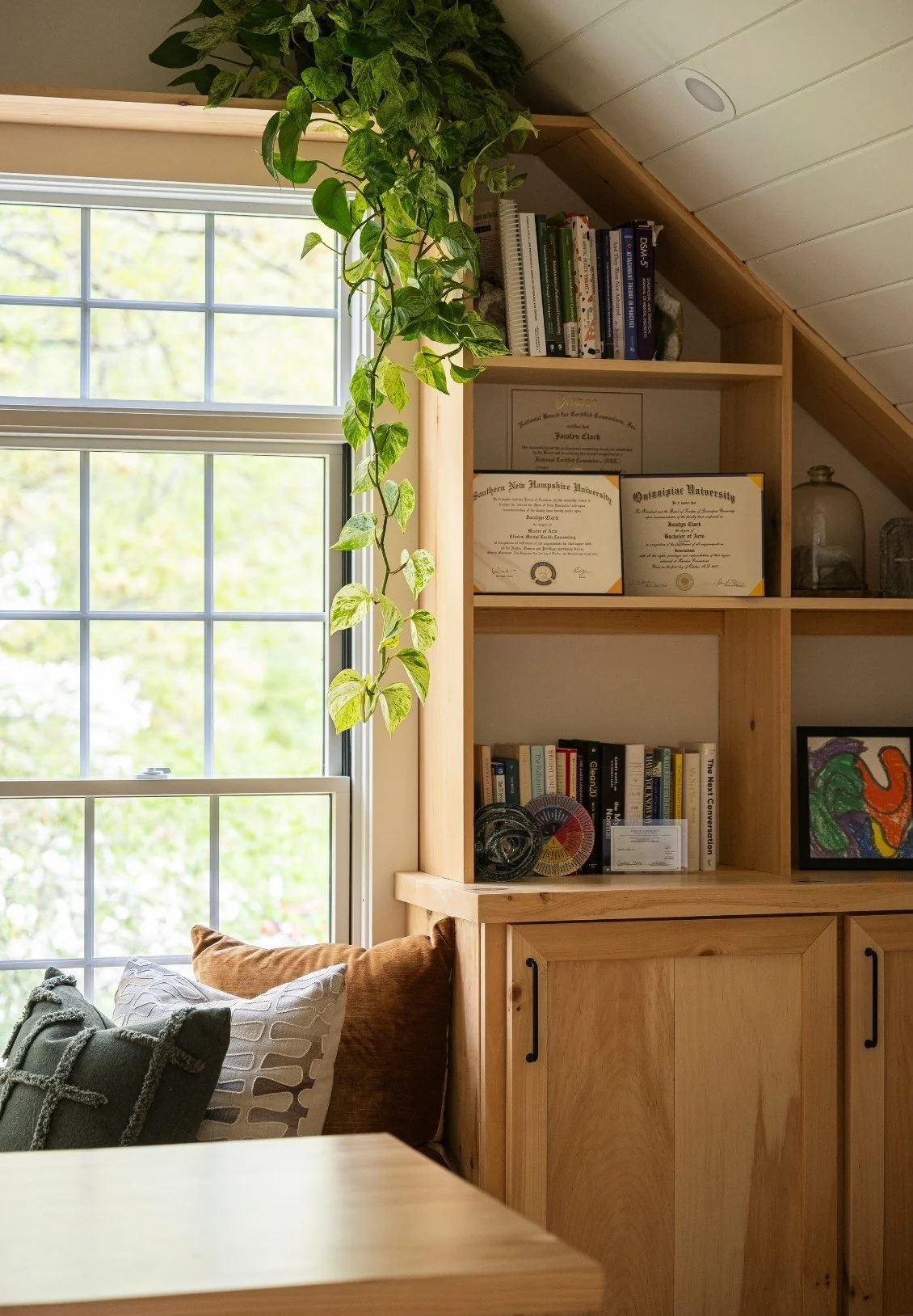 A cozy corner of a room with a wooden bookshelf filled with books and framed certificates. A large window with sunlight streaming in, and a green leafy plant draping down from above. A section of sofa with pillows is visible at the bottom left.