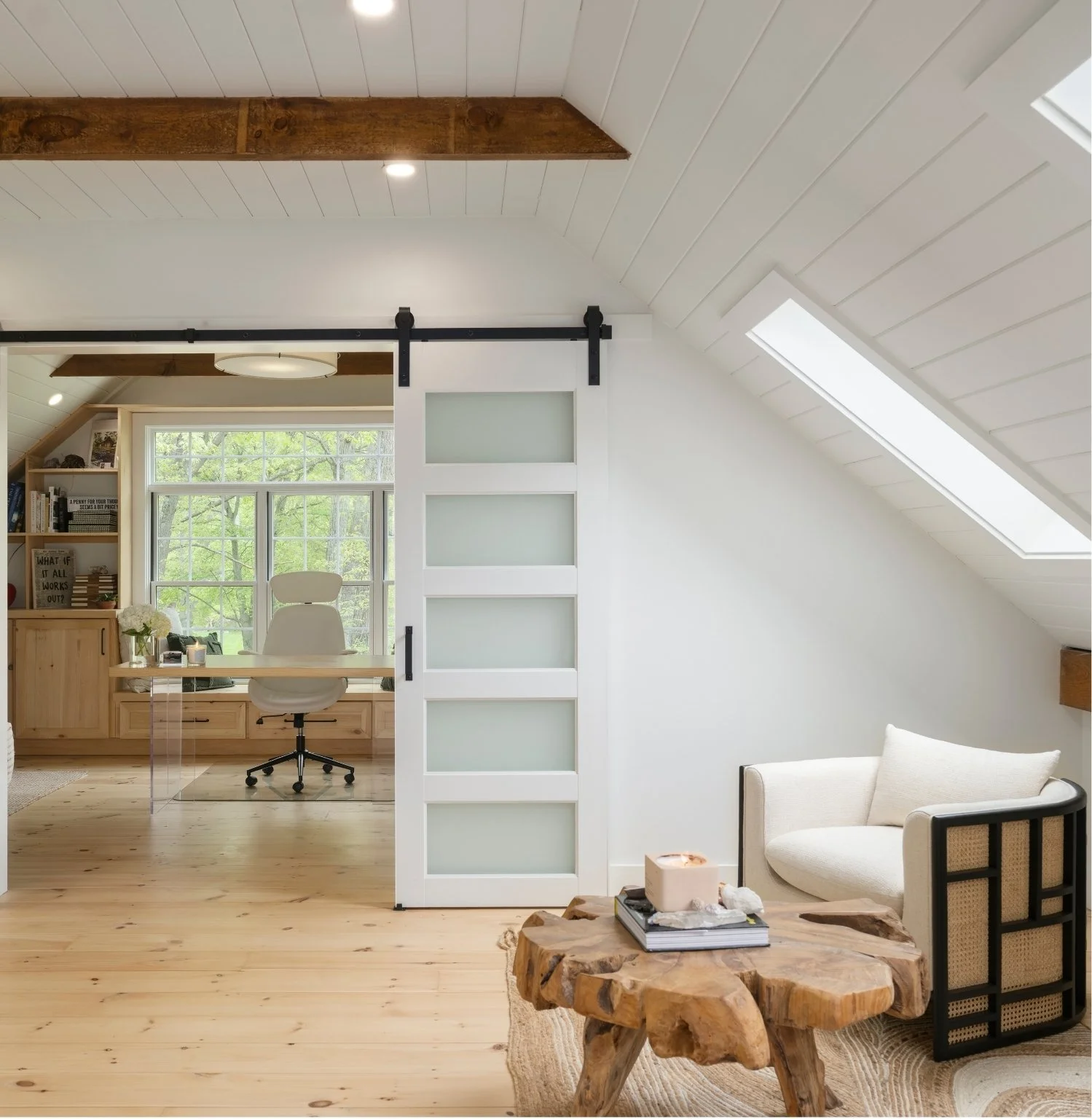 A cozy room with a beige armchair and a rustic wooden coffee table. A white sliding barn door with frosted glass panels separates the room from a sunlit home office with a window and greenery outside. The room has vaulted ceilings with white shiplap and skylights, and wood flooring.