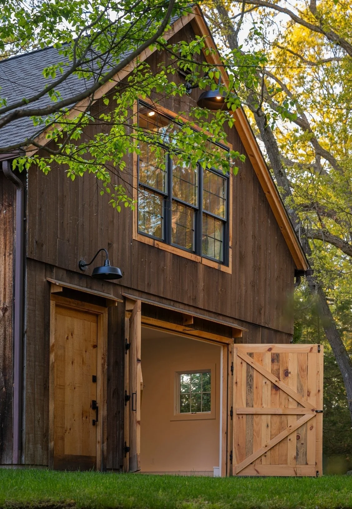 A rustic barn with a wooden exterior, large black-framed window, open double doors, and surrounded by trees with green and yellow leaves.