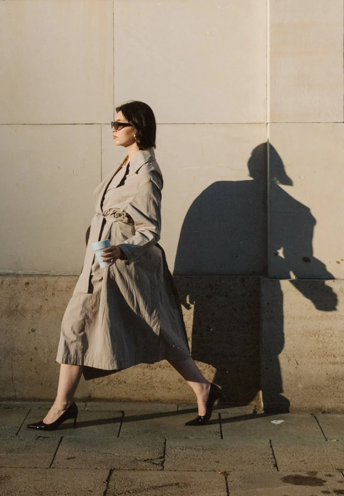 A woman walking on the sidewalk at night, holding a cup, wearing a beige trench coat, black heels, sunglasses, and big earrings. Her shadow is cast on a wall behind her.