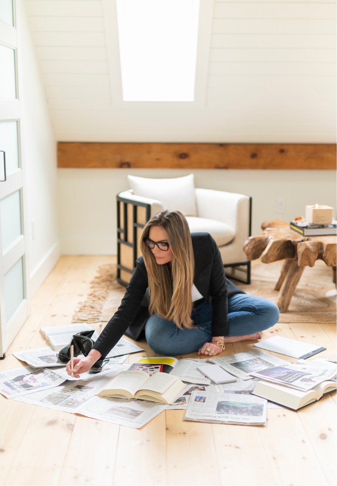 A woman with long blonde hair, wearing glasses, a black blazer, and jeans, sitting on the wooden floor of a bright room amidst scattered newspapers, open books, and notebooks, with a white armchair and rustic wooden table in the background.