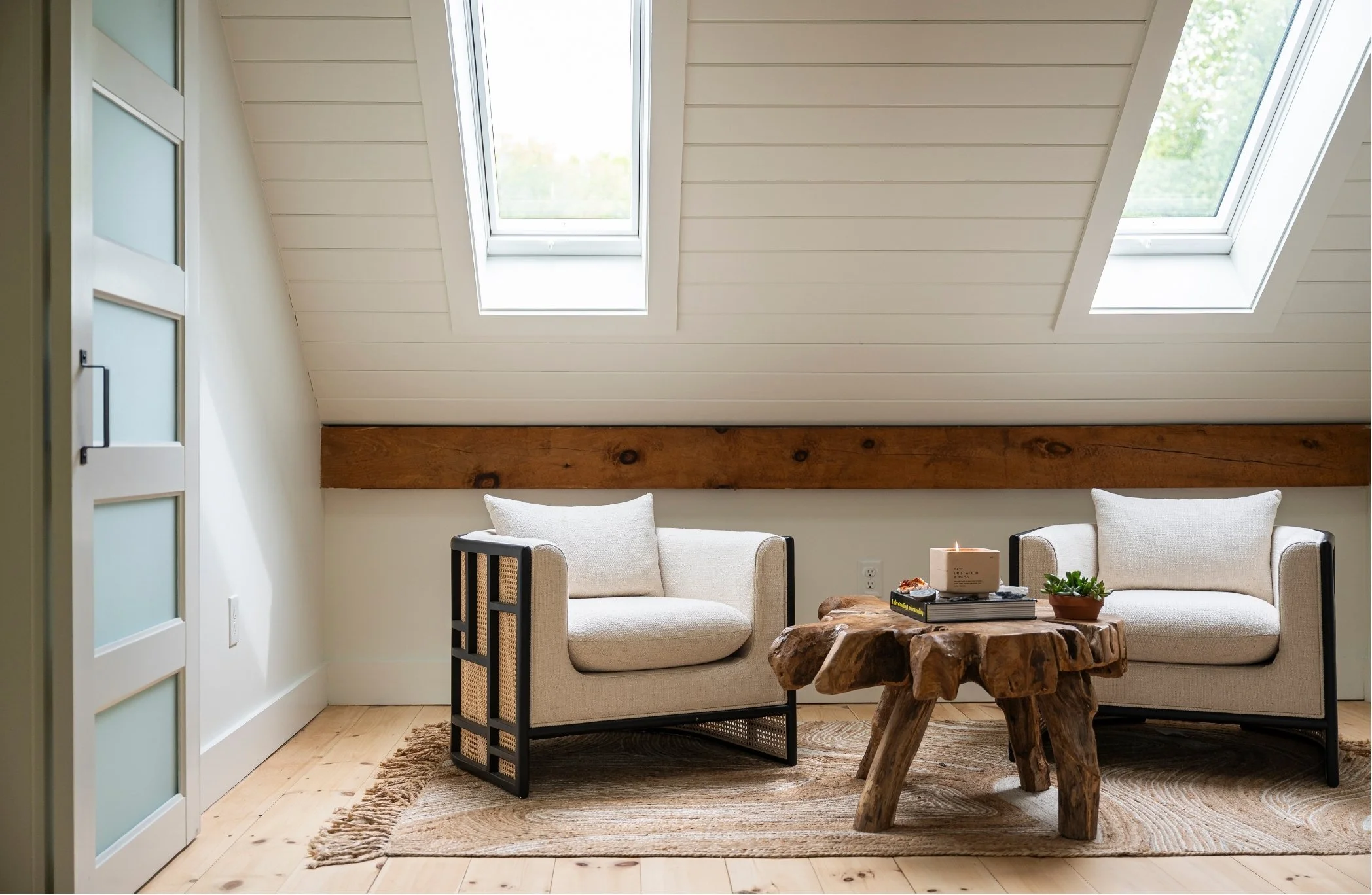 A cozy living room with a sloped ceiling featuring two skylight windows and white shiplap walls. There are two off-white sofas, a rustic wooden coffee table, and a small slide closet door. A small potted plant and a candle are on the coffee table, and a woven rug covers the wooden floor.