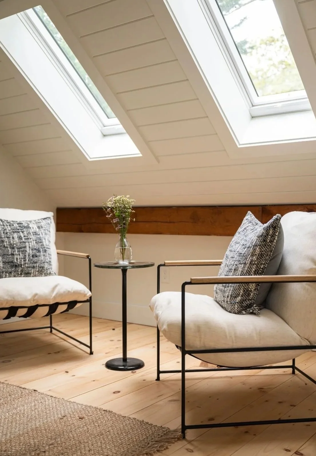 A cozy attic room with a sloped ceiling and two skylight windows, featuring two chairs with cushions, a small round table with a vase of flowers, and a natural wood floor.