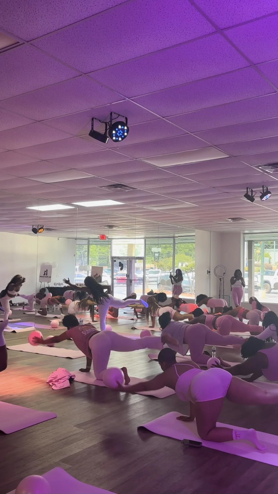 Group of people participating in a yoga class in a brightly lit studio with purple lighting. They are in various yoga poses on mats, with some individuals stretching and others in different positions of physical exercise.
