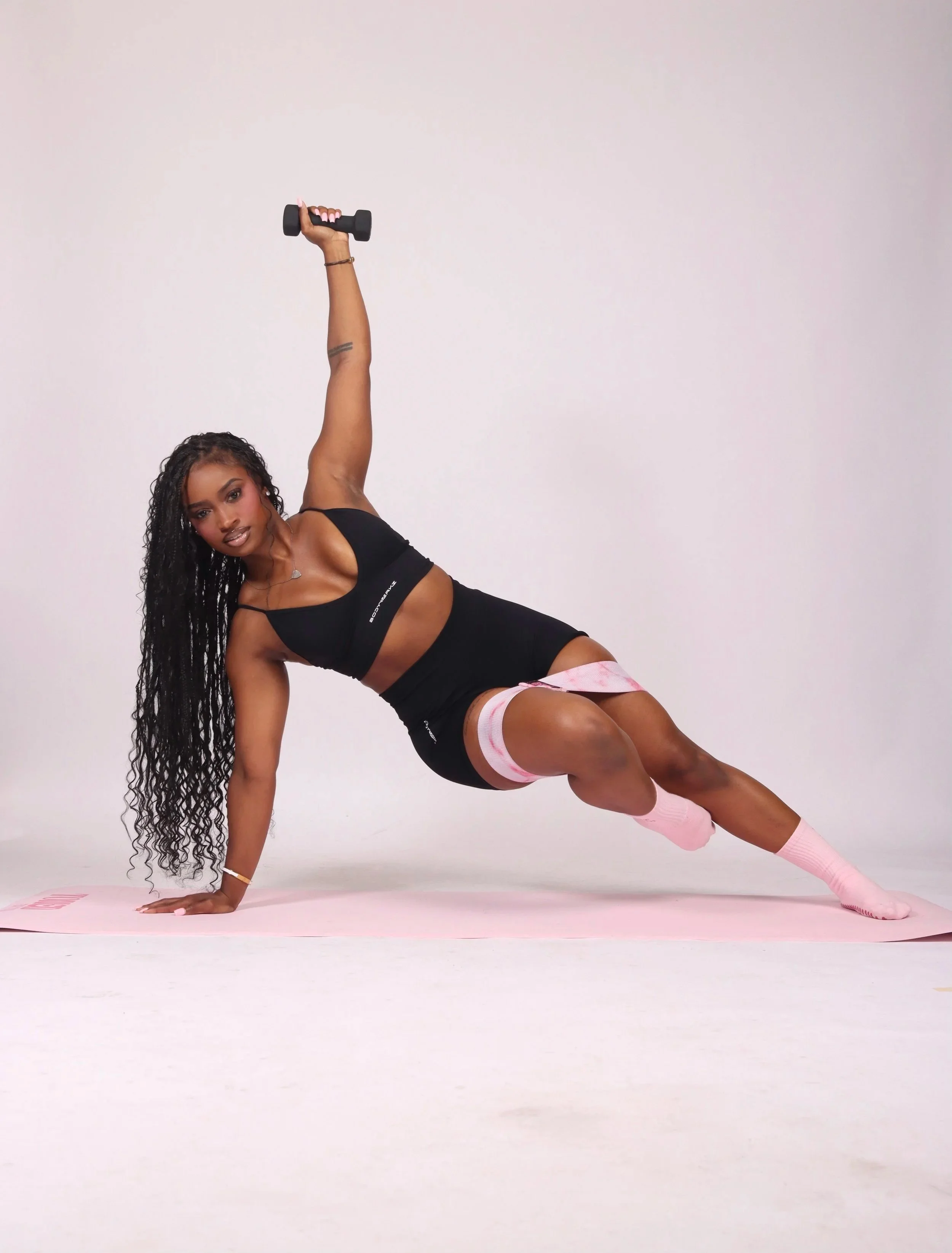 A woman with long curly hair and medium skin tone exercising on a pink yoga mat in a studio with a plain light pink background. She is wearing a black sports bra, matching shorts, pink striped socks, and holding a black dumbbell in her right hand, extended upwards while in a side plank pose.