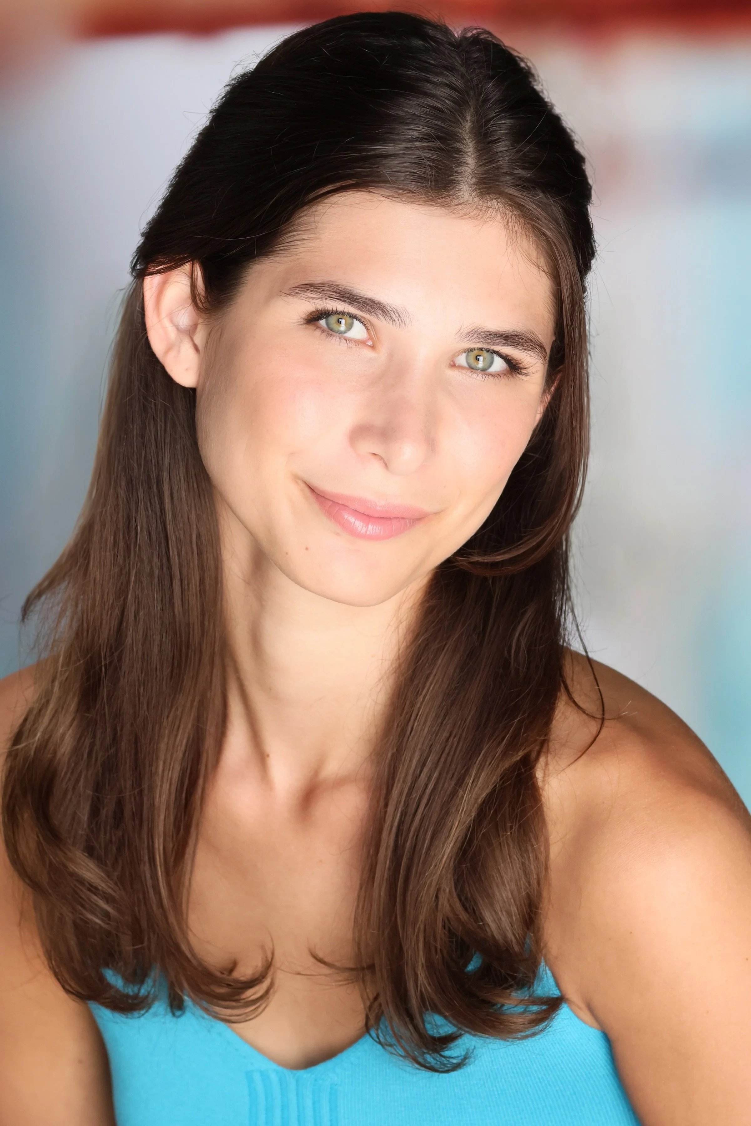 Close-up of a young woman with long brown hair, green eyes, and a gentle smile, wearing a turquoise top, against a blurred multicolored background.