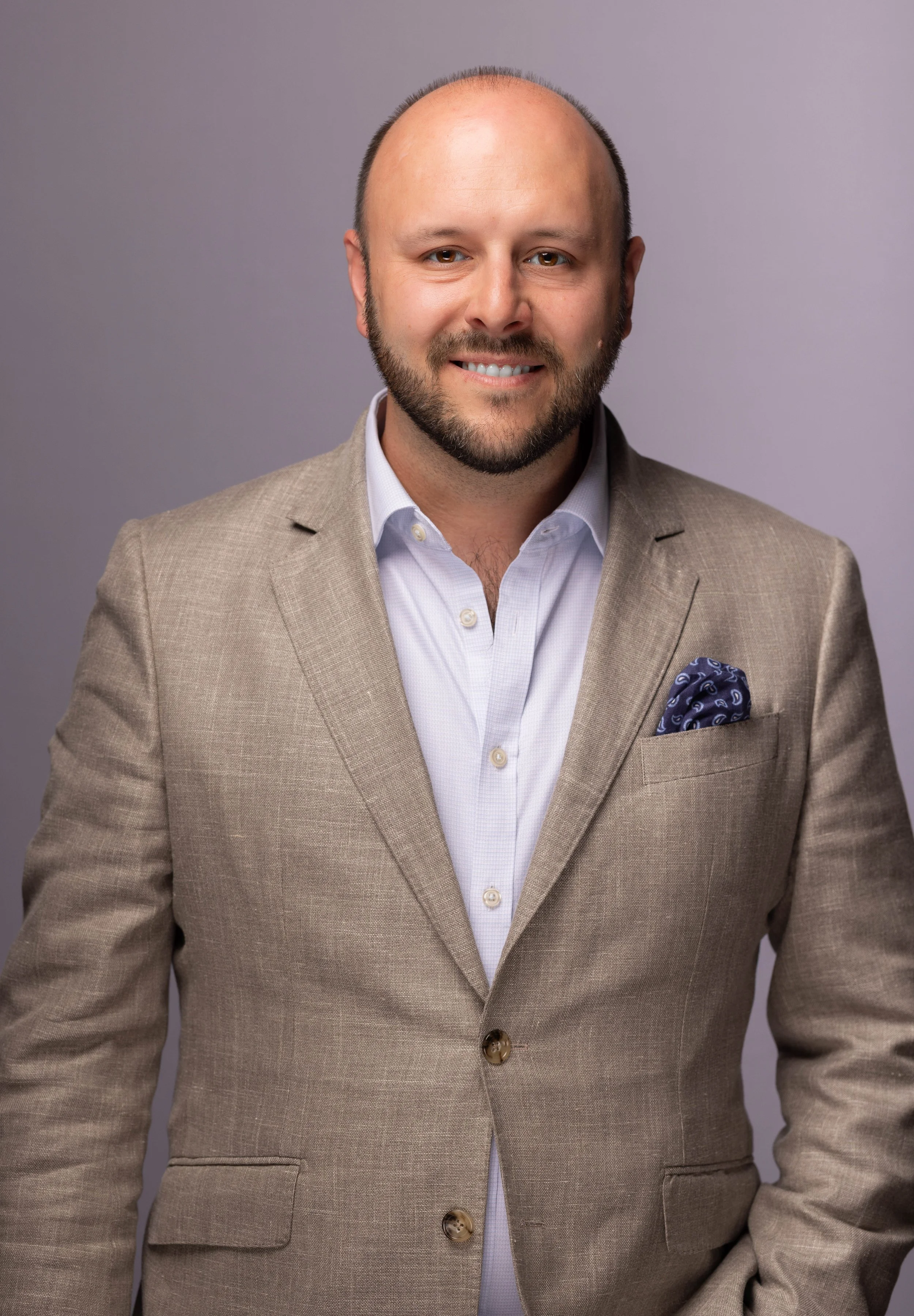 A headshot of a smiling man with light skin, a beard, and a receding hairline, dressed in a beige suit jacket and a light purple dress shirt, against a plain gray background.