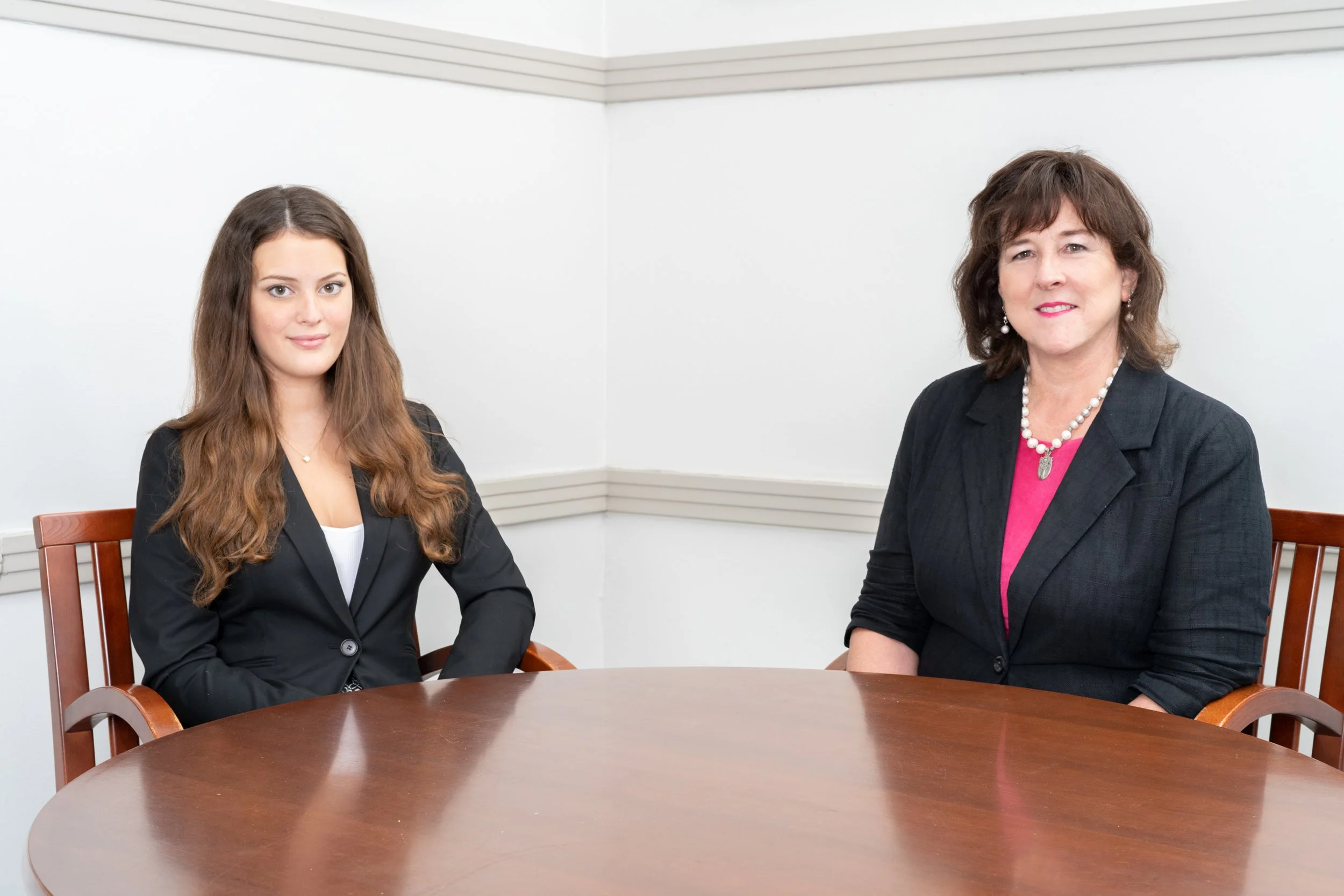Two women sitting at a round wooden table in a meeting room with white walls and chair backs. The woman on the left has long, wavy brown hair and is wearing a black blazer. The woman on the right has short, curly brown hair and is dressed in a dark blazer with a pink top and jewelry.