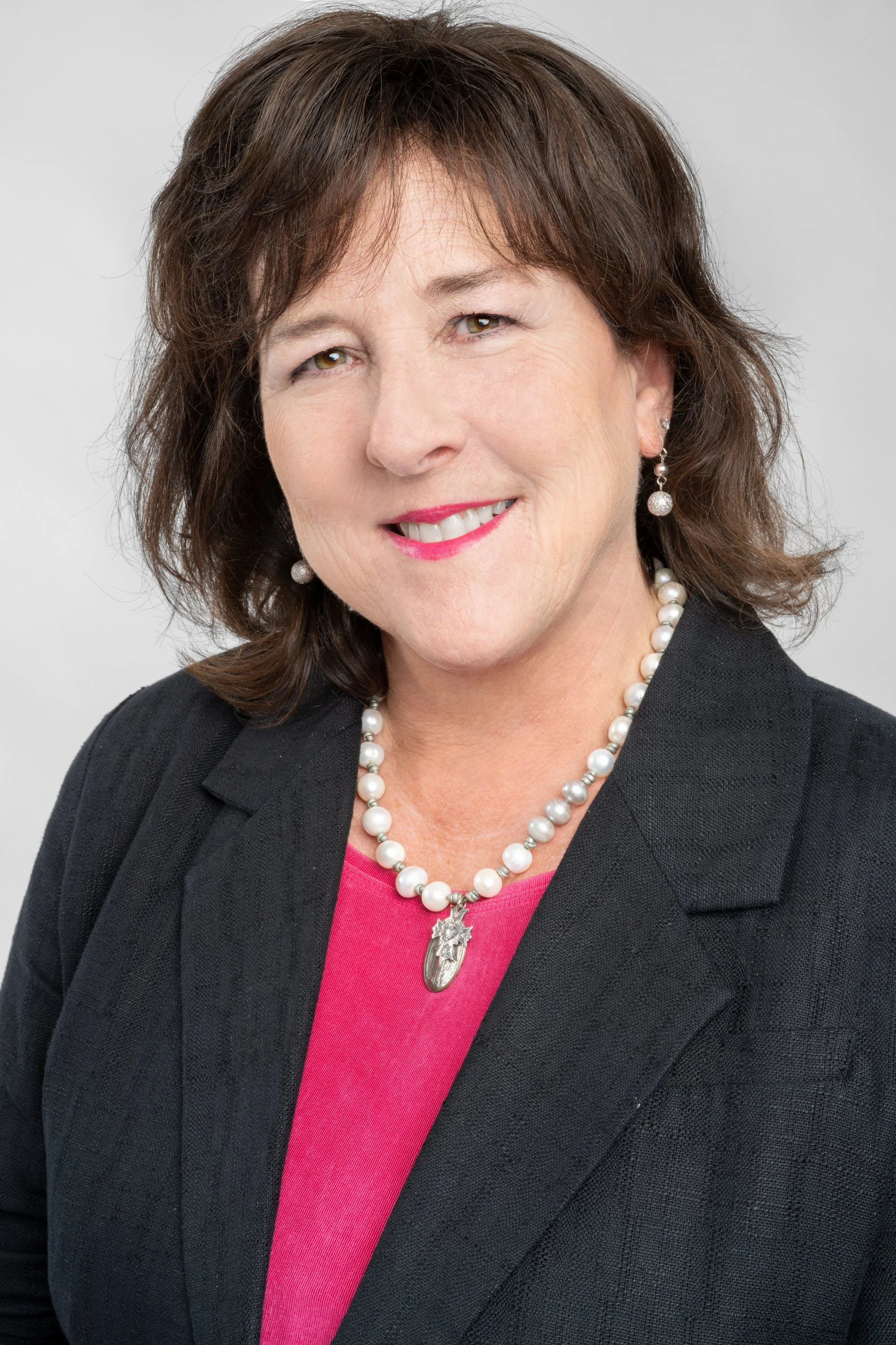 A middle-aged woman with shoulder-length brown hair, wearing pearl earrings, a pearl necklace, a black blazer, and a pink top, smiling at the camera against a plain gray background.