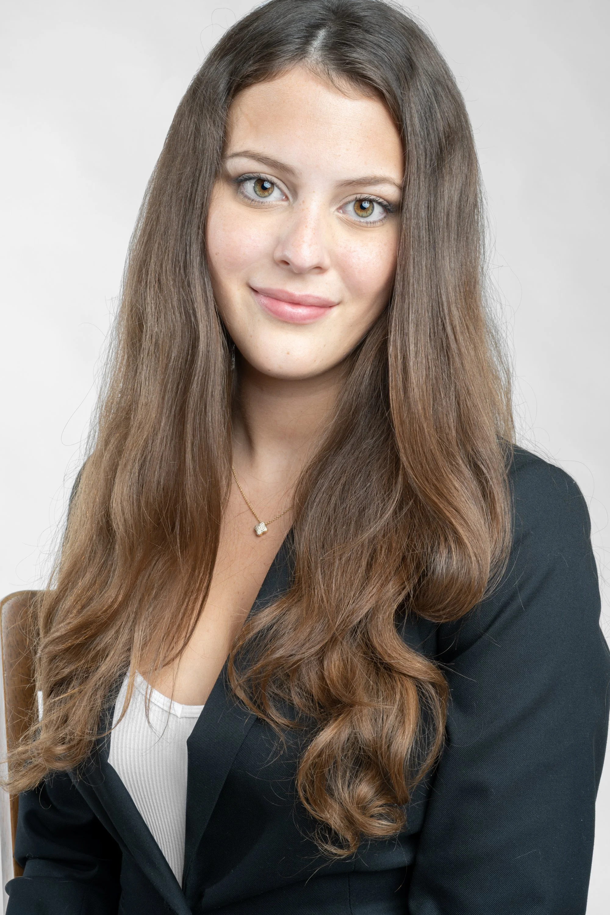 A woman with long brown hair, wearing a black blazer and a white top, with a gold necklace, smiling at the camera against a plain light background.
