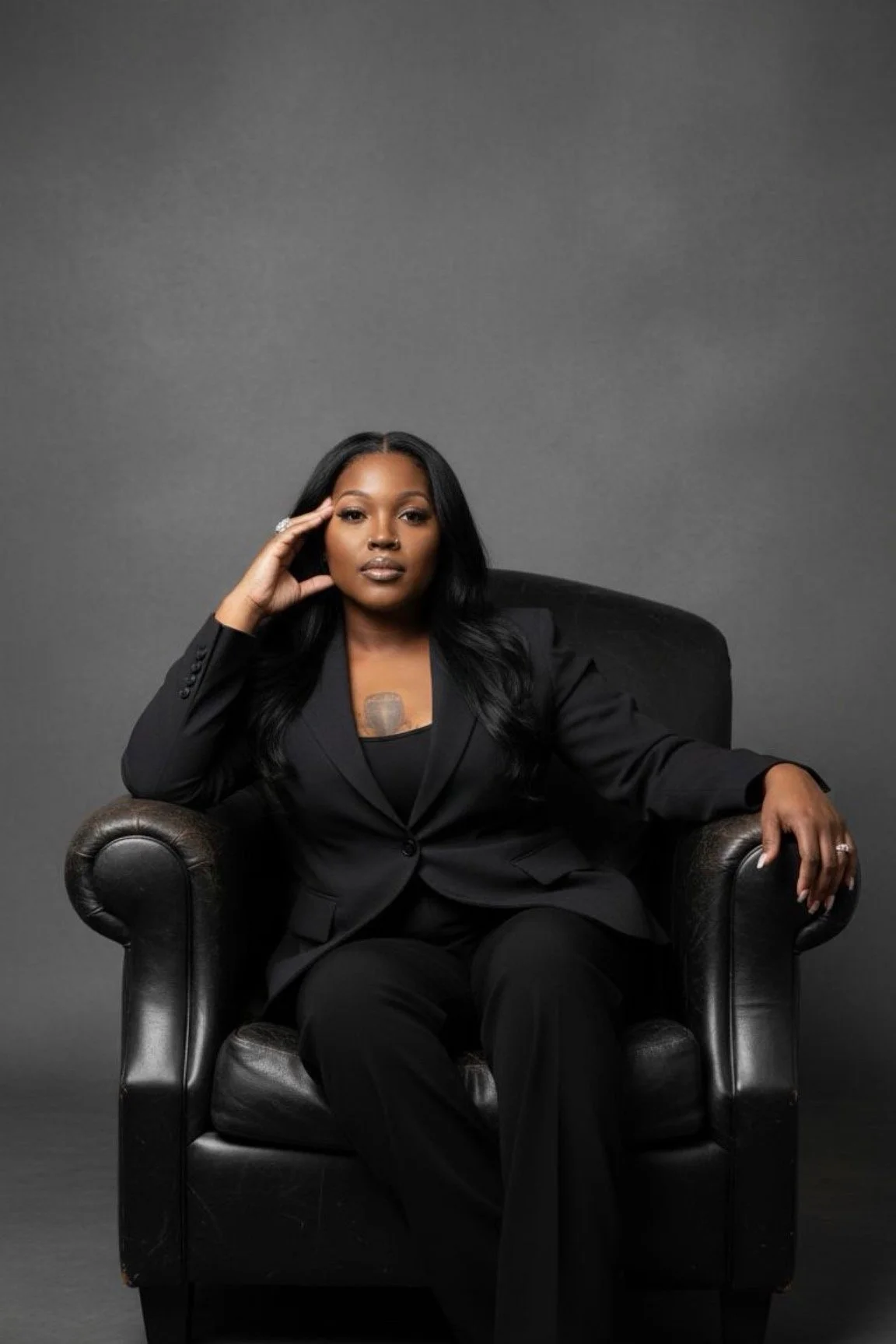 A woman in a black suit sitting on a black leather armchair against a plain gray background. She has long black hair and is posing thoughtfully with her right hand on her temple and her left hand resting on the arm of the chair.