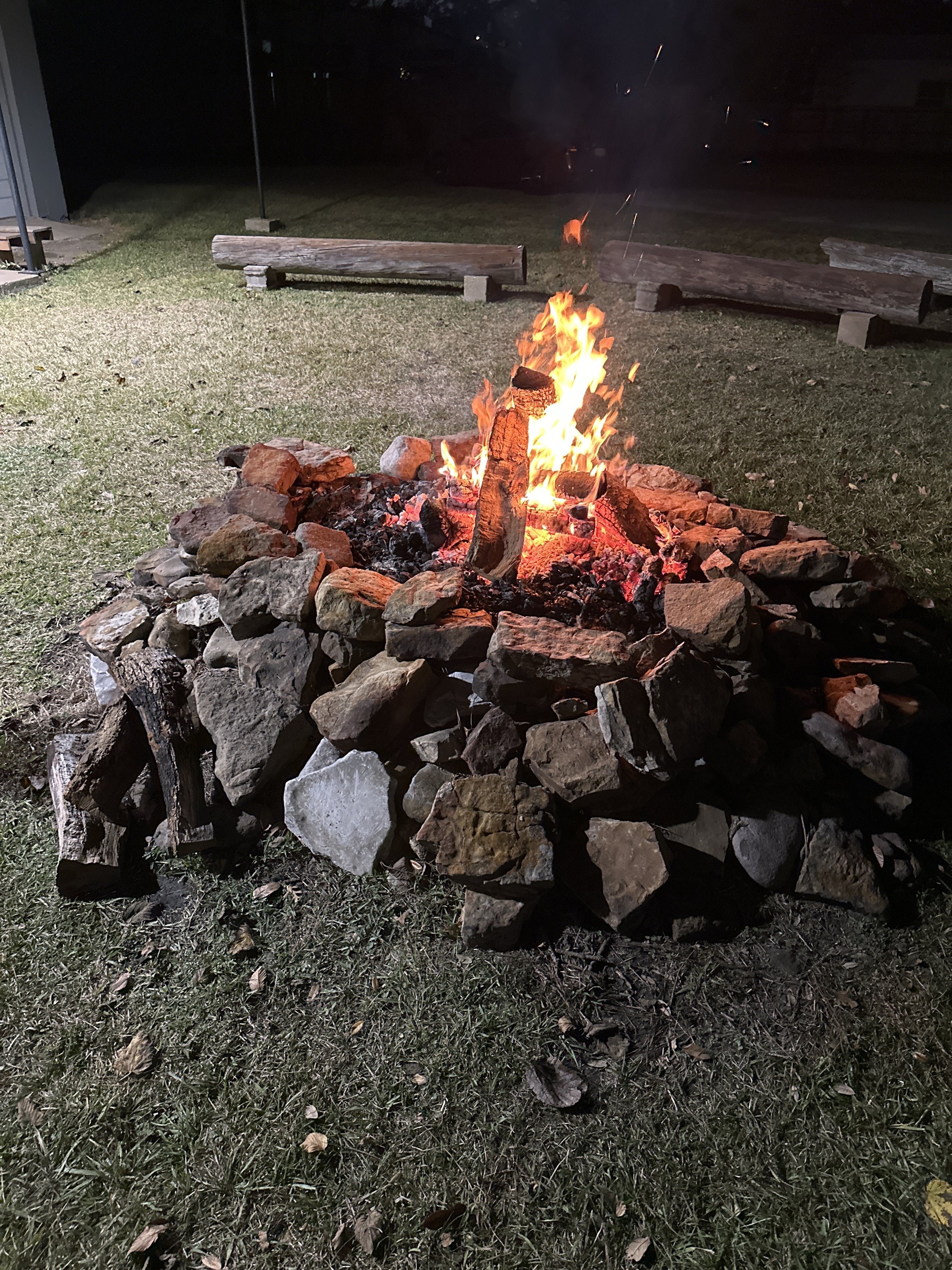 A campfire surrounded by rocks on grass at night, with wooden benches in the background.