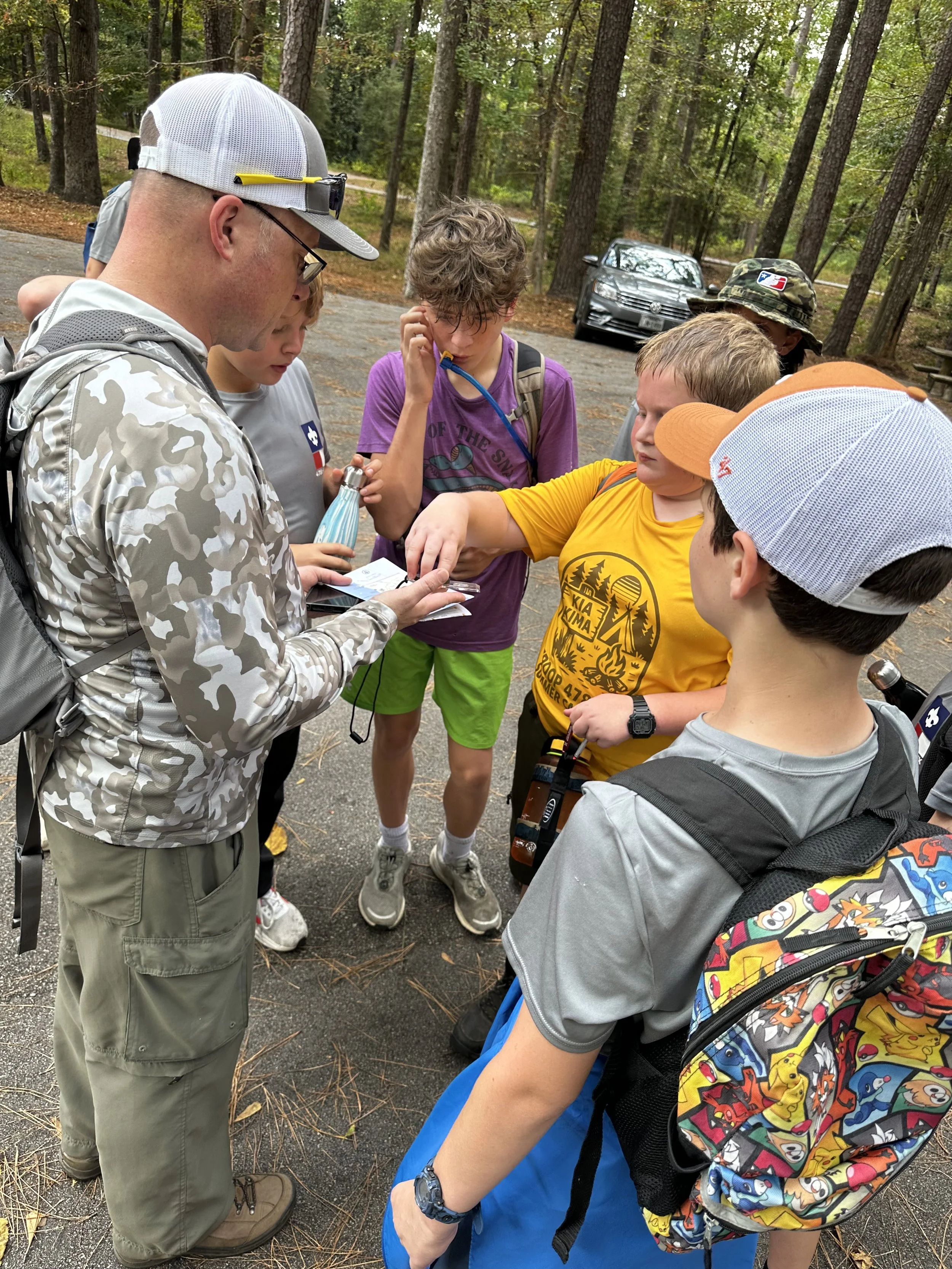 A group of children and an adult gathering outdoors in a wooded area, looking at a map or phone.