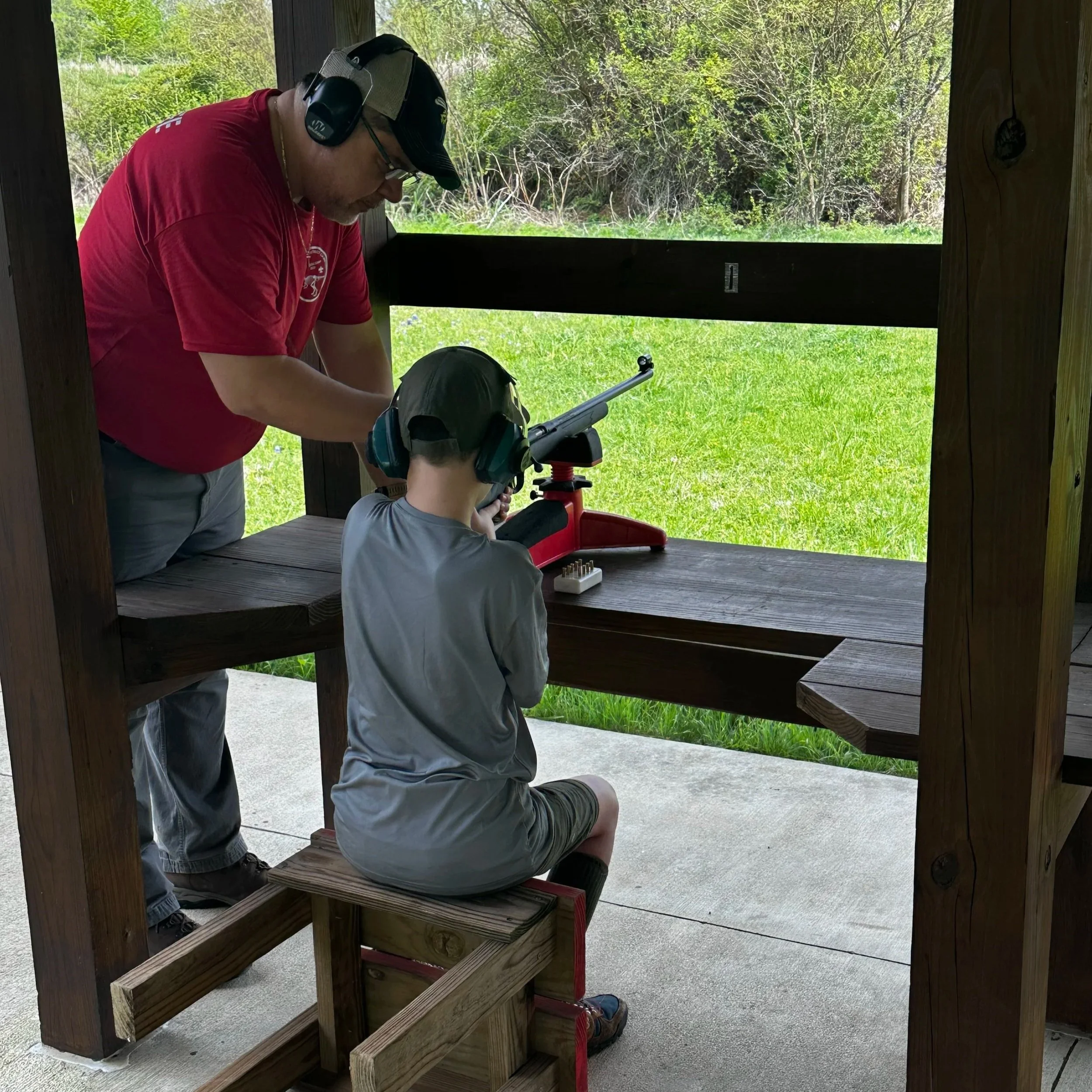 An adult man with a hat, glasses, and headphones, and a boy with ear protection, are at a shooting range with a rifle on a rest. They are inside a wooden shelter with green grass and trees outside.