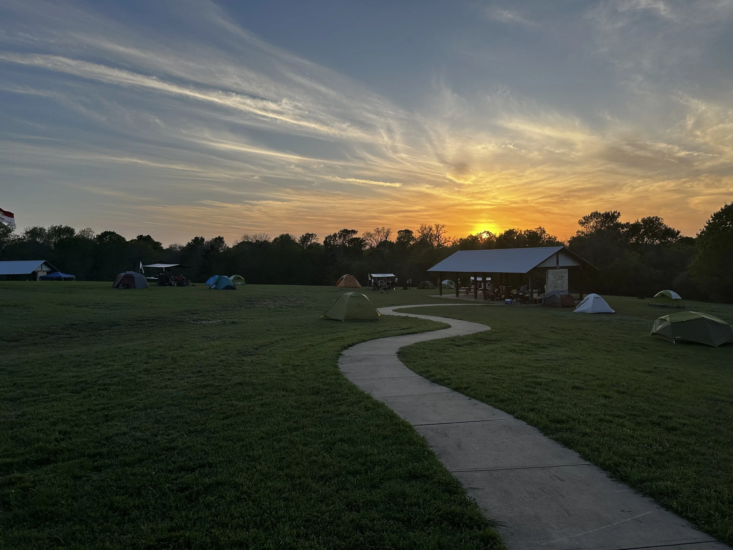 Sunset over a grassy outdoor campground area with tents, a pavilion, trees, and a paved walking path.