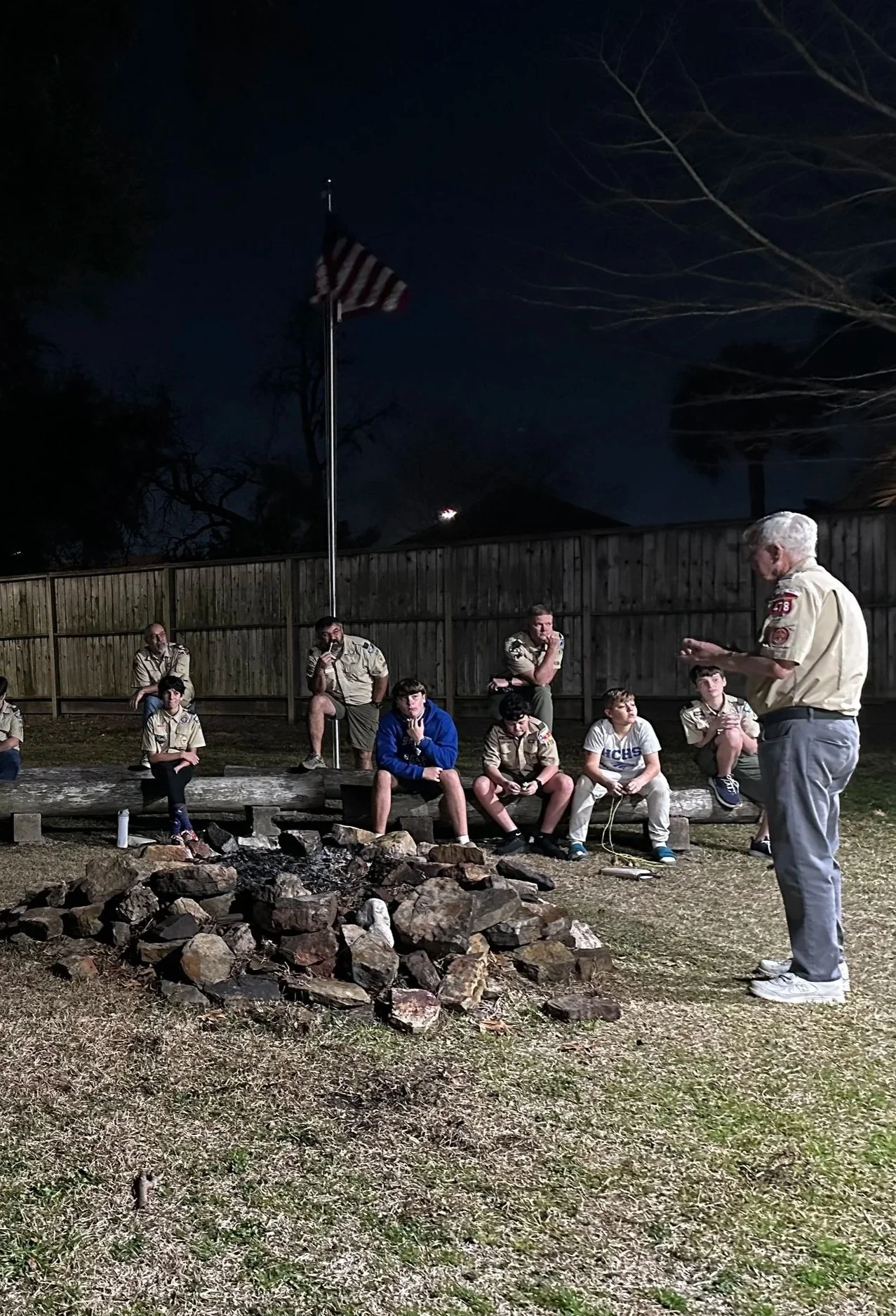 A group of Boy Scouts sitting and standing around a campfire pit at night, listening to an adult leader who is reading or speaking, with a wooden fence and American flag in the background.