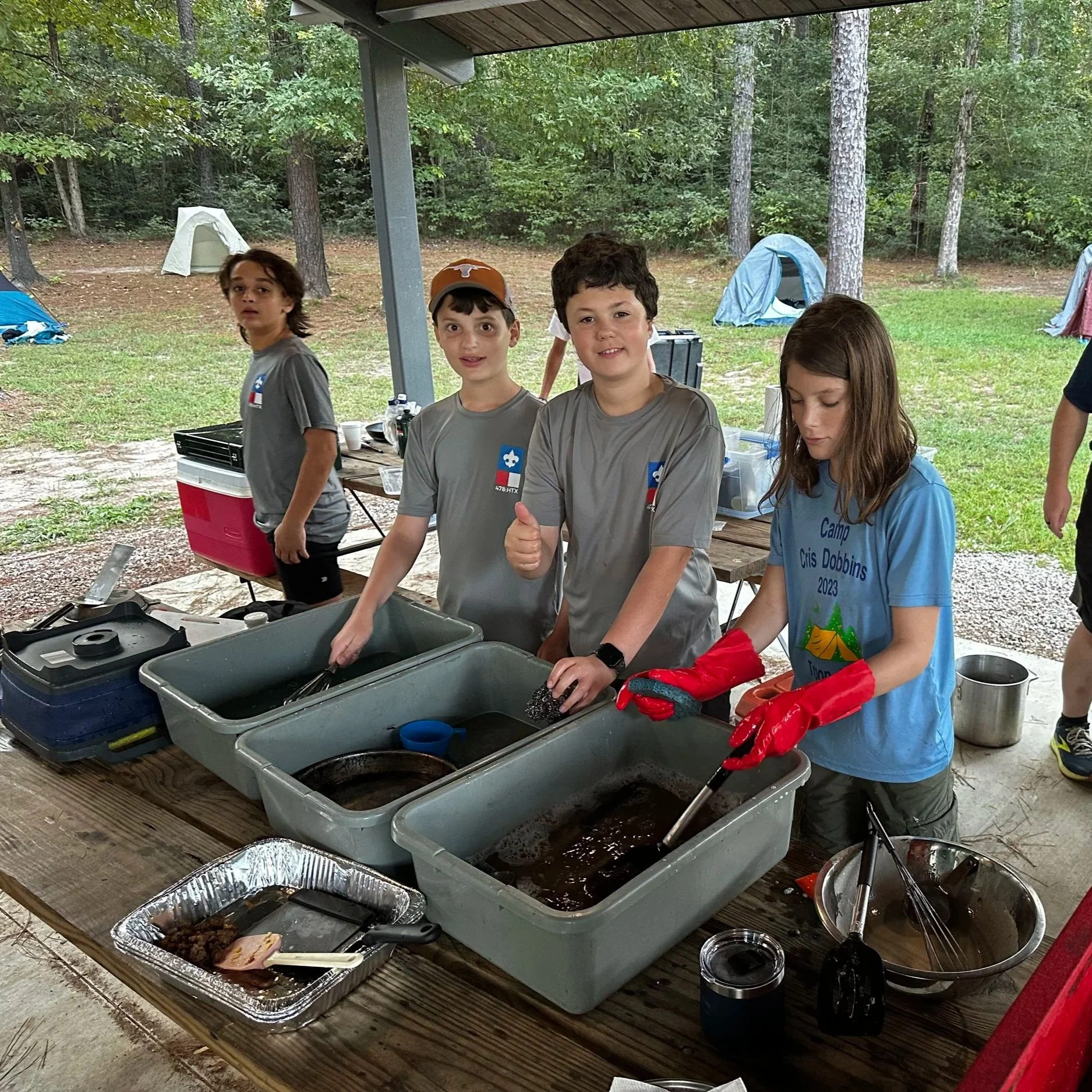 A group of children at a summer camp standing around a table with food containers, some giving thumbs up and others looking at the camera, outdoors in a wooded area with tents in the background.