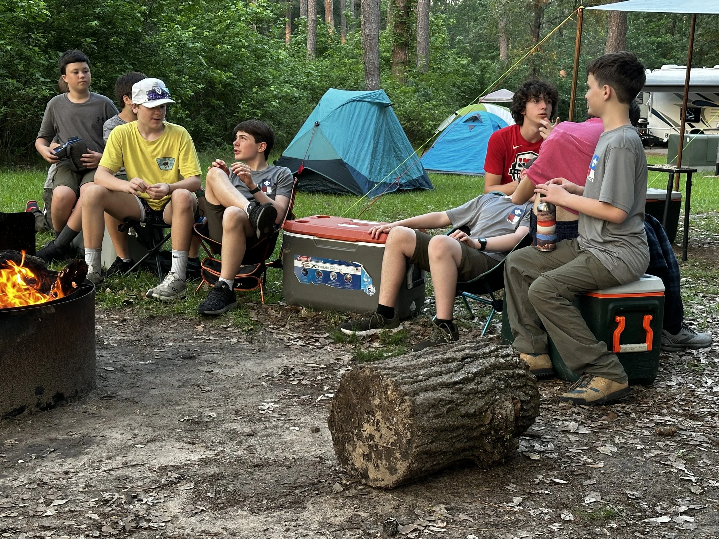 Group of boys sitting around a campfire during a camping trip in the woods, with tents and camping gear around them.
