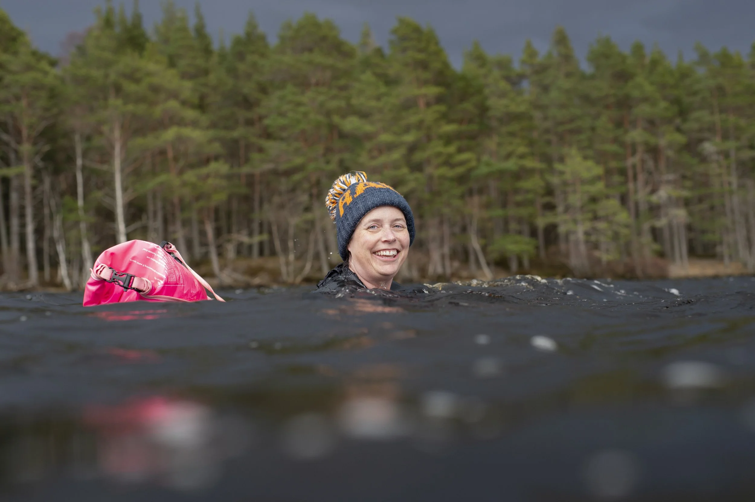 Smiling woman swims in a lake wearing a wooley hat and with woodland in the background