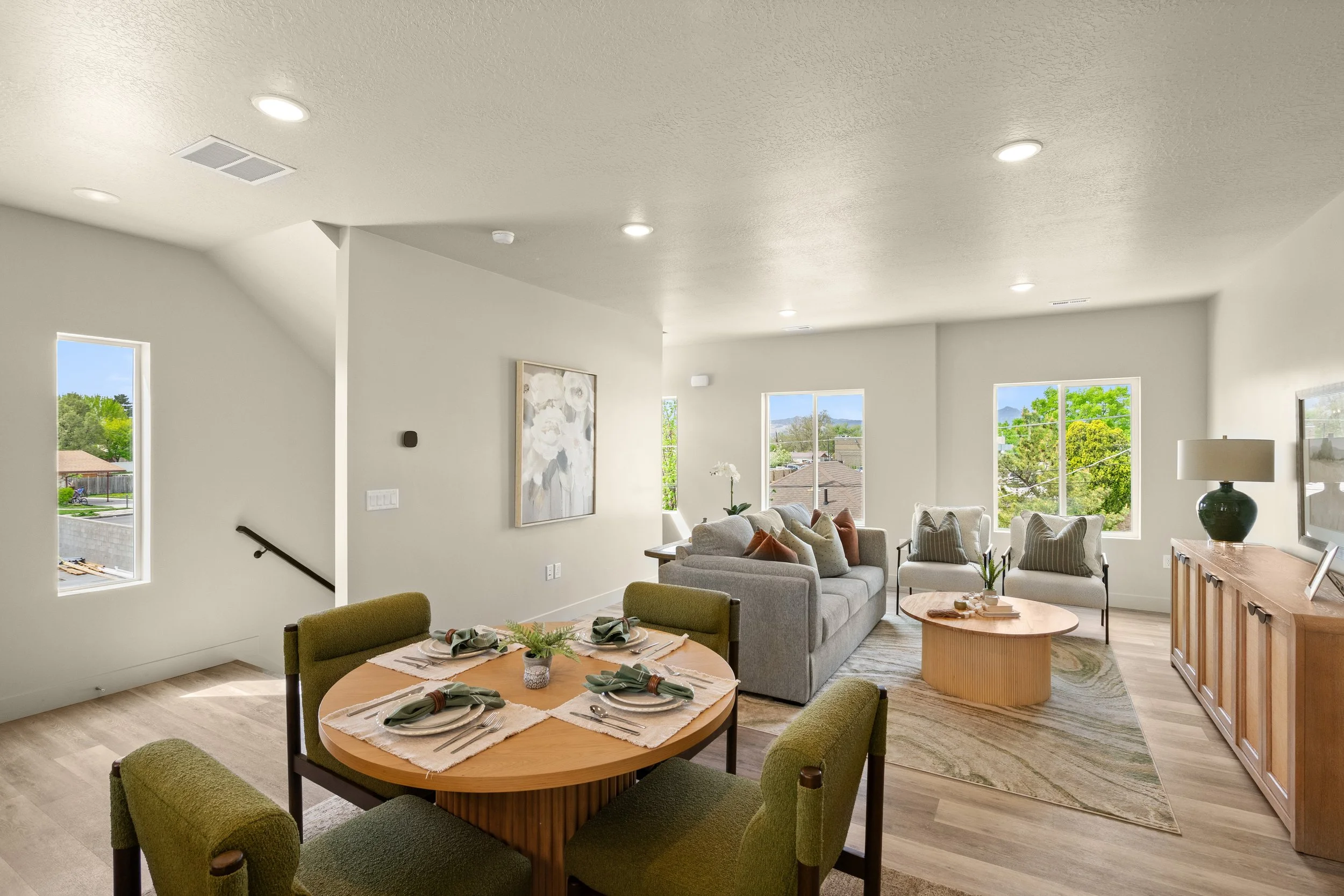 Bright living room with large windows, beige sofa, chairs, and a dining table set with napkins and greenery.