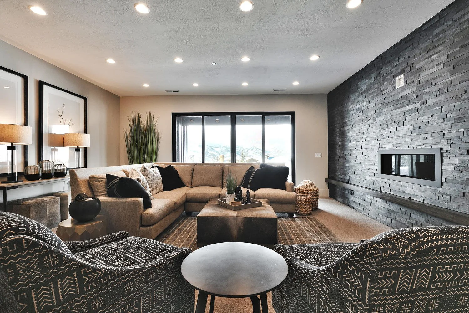 Modern living room with a beige sectional sofa, black and beige patterned armchairs, a dark fireplace on a textured gray brick wall, large windows in the background, decorative items like lamps, vases, and a tray with small objects on a central coffe