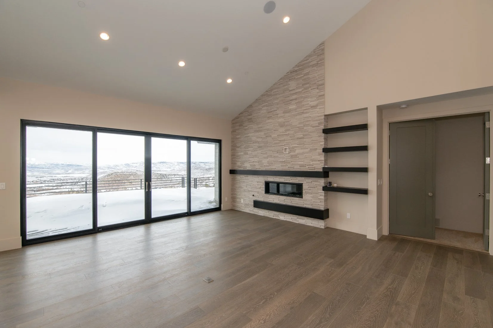 A spacious living room with large sliding glass doors opening to a snow-covered balcony with mountain views. The room features a modern stone accent wall with a fireplace and black floating shelves. Light-colored hardwood floors and a beige wall comp