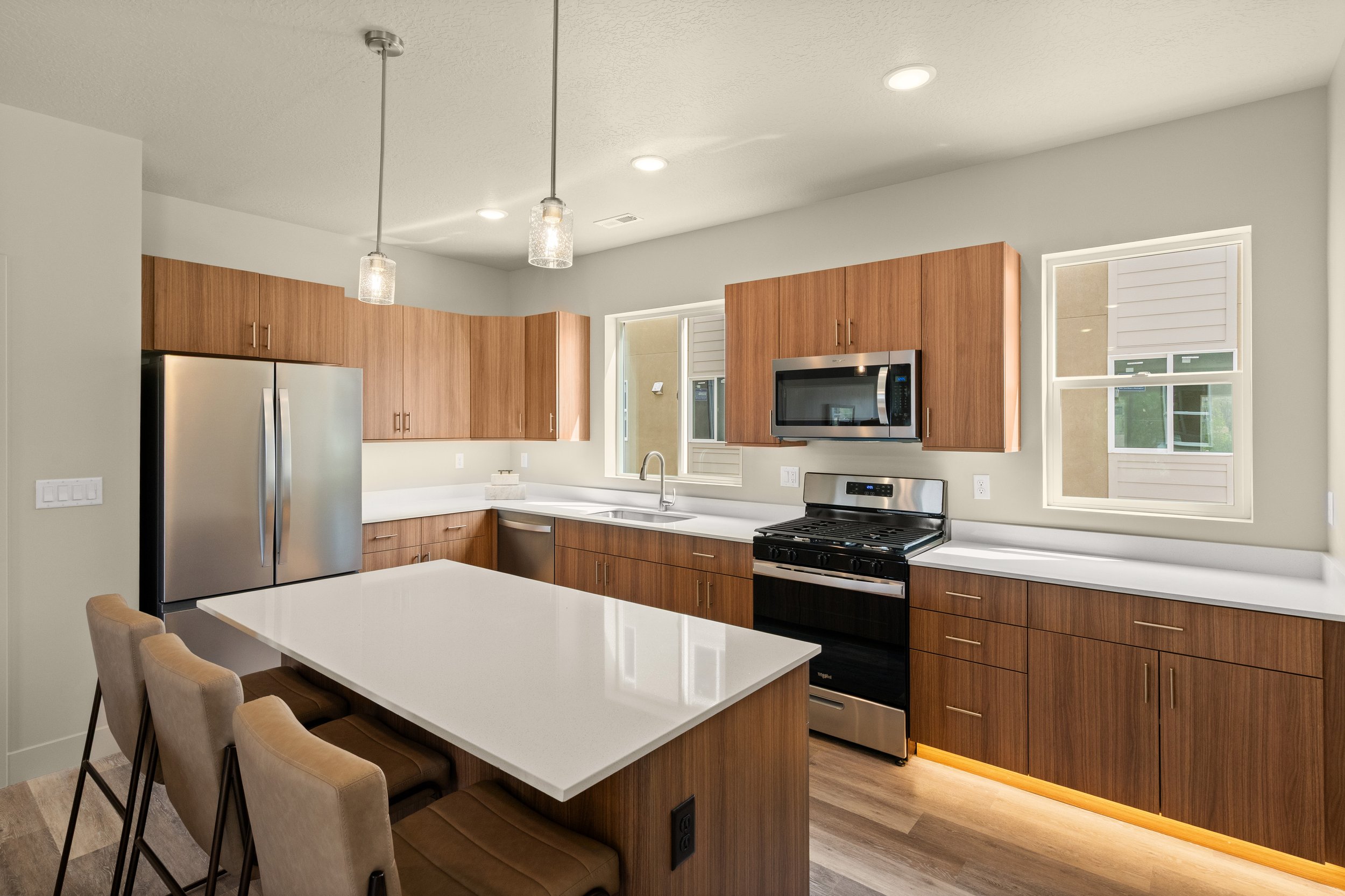 Modern kitchen with wooden cabinets, stainless steel appliances including a refrigerator, microwave, and oven, a white island with seating, and windows providing natural light.