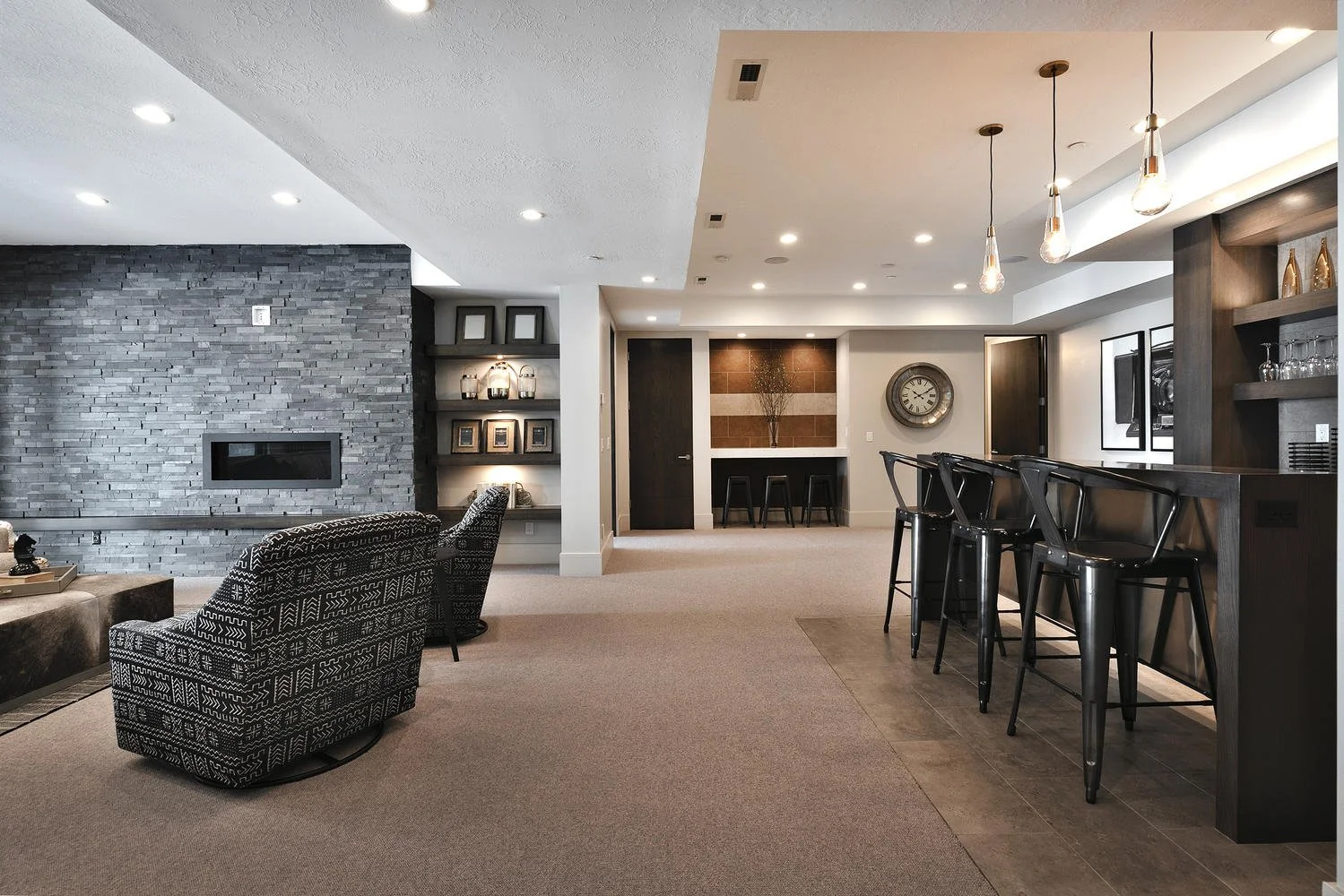 Modern living room with a gray stone wall fireplace, patterned armchairs, a bar area with high black chairs, and minimalist decor.
