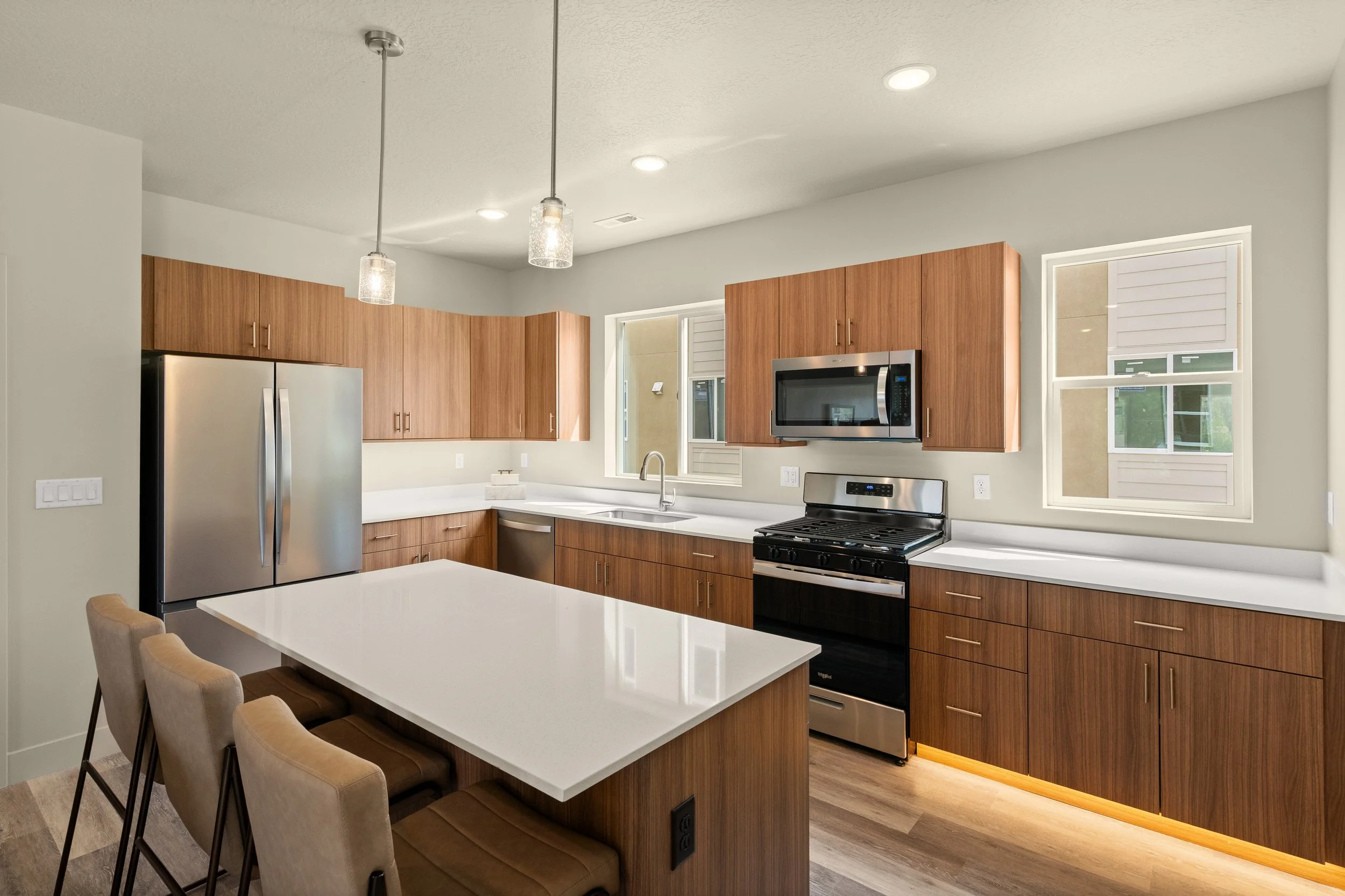 Modern kitchen with wooden cabinets, a white countertop, stainless steel appliances including a refrigerator and oven, a microwave above the oven, and a double sink under a large window. There is a white island with seating and pendant lights hanging from the ceiling.