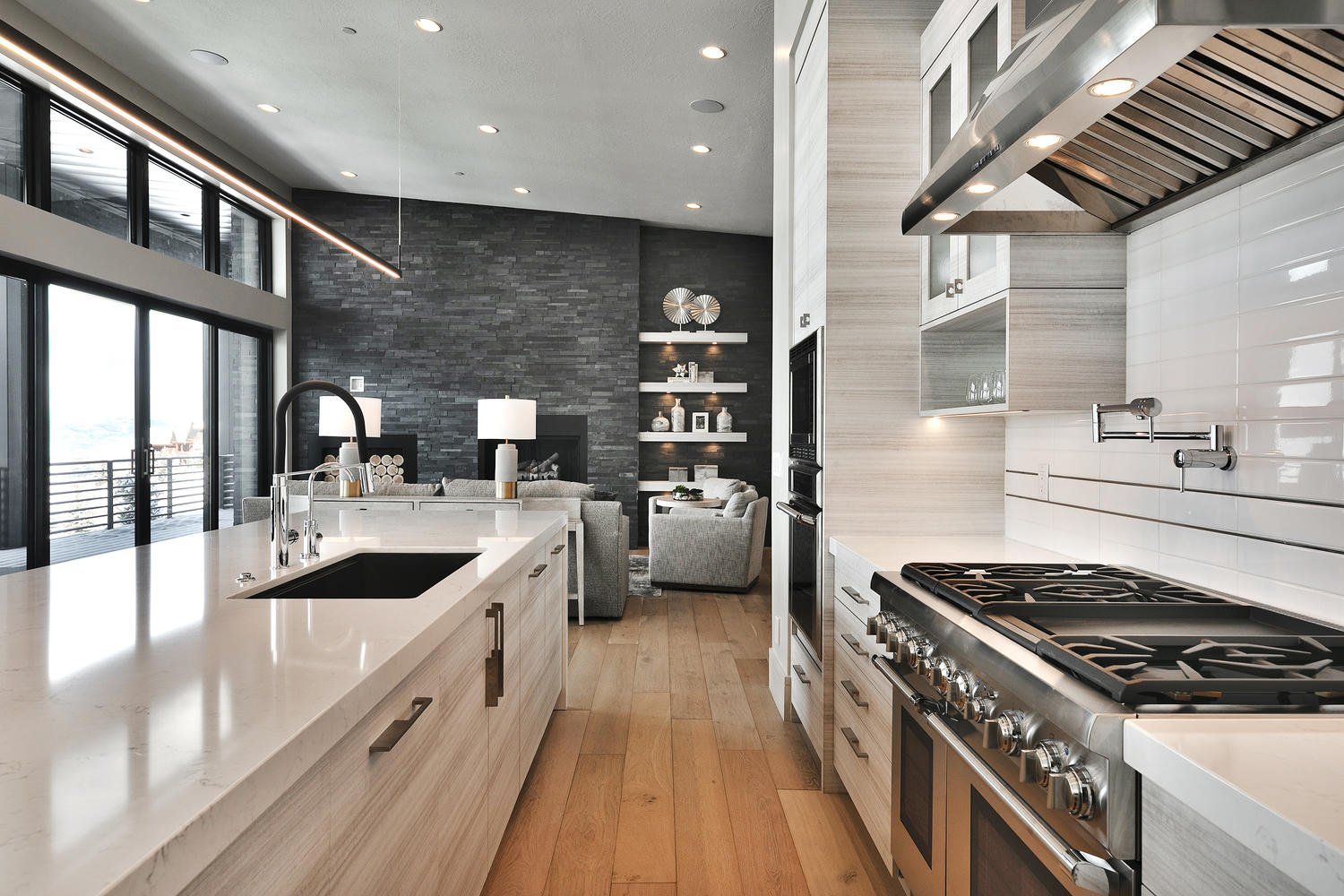Modern open-concept kitchen with white island, black sink, and stainless steel appliances, with a living area featuring a dark accent wall, gray seating, and decorative shelving.