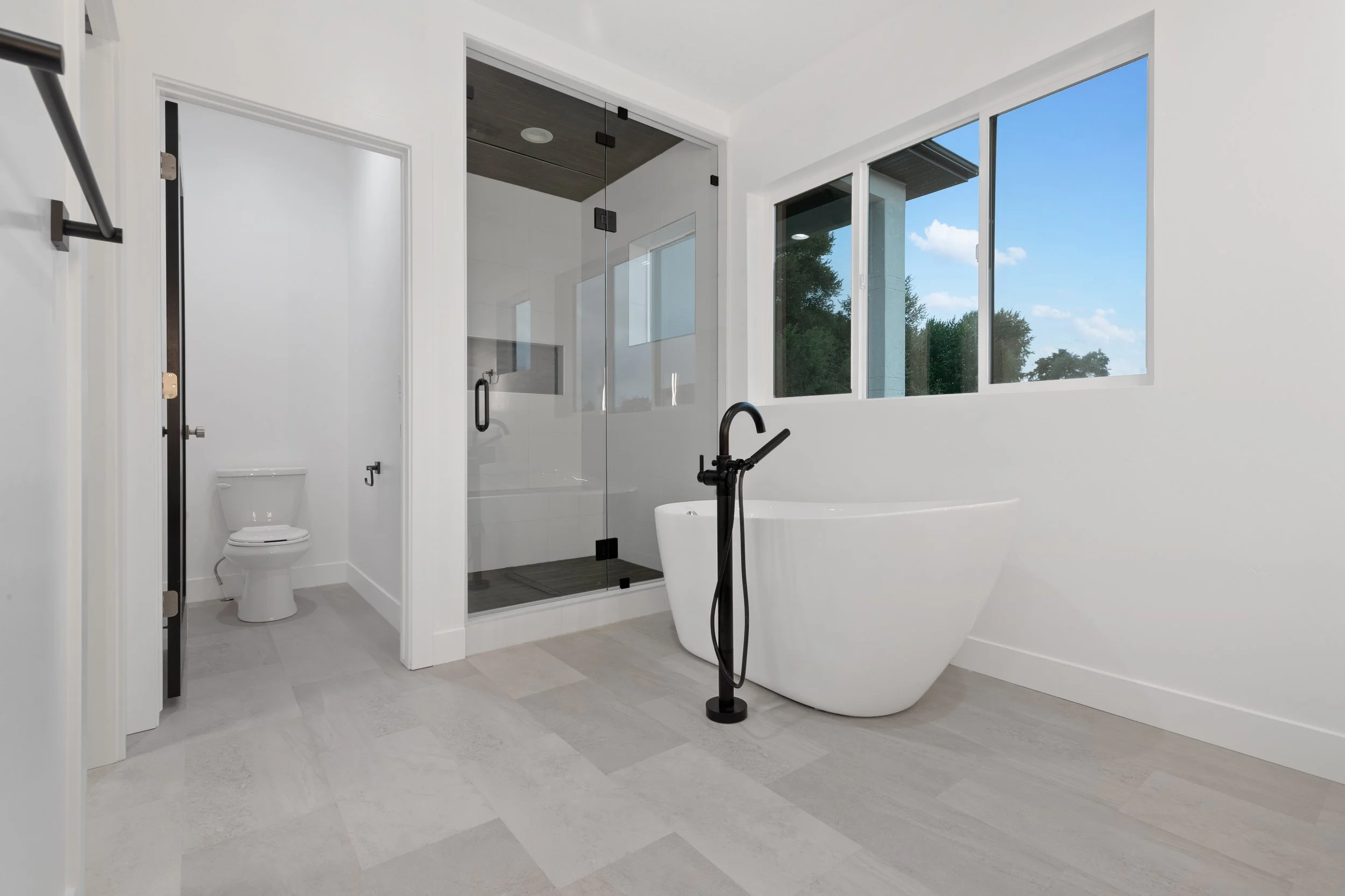 Modern bathroom with walk-in shower, freestanding tub, large windows, white walls, and light gray flooring.