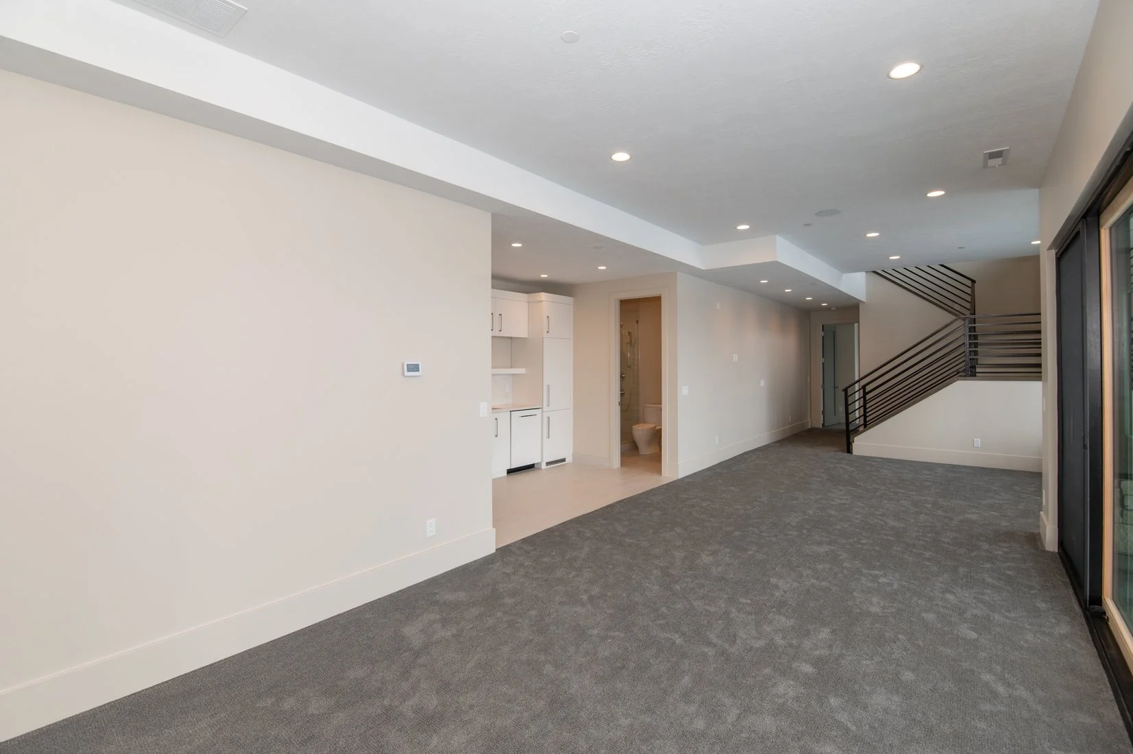 Empty living room with beige walls, gray carpet, built-in kitchenette, a small bathroom, and a staircase with black railings leading upstairs.