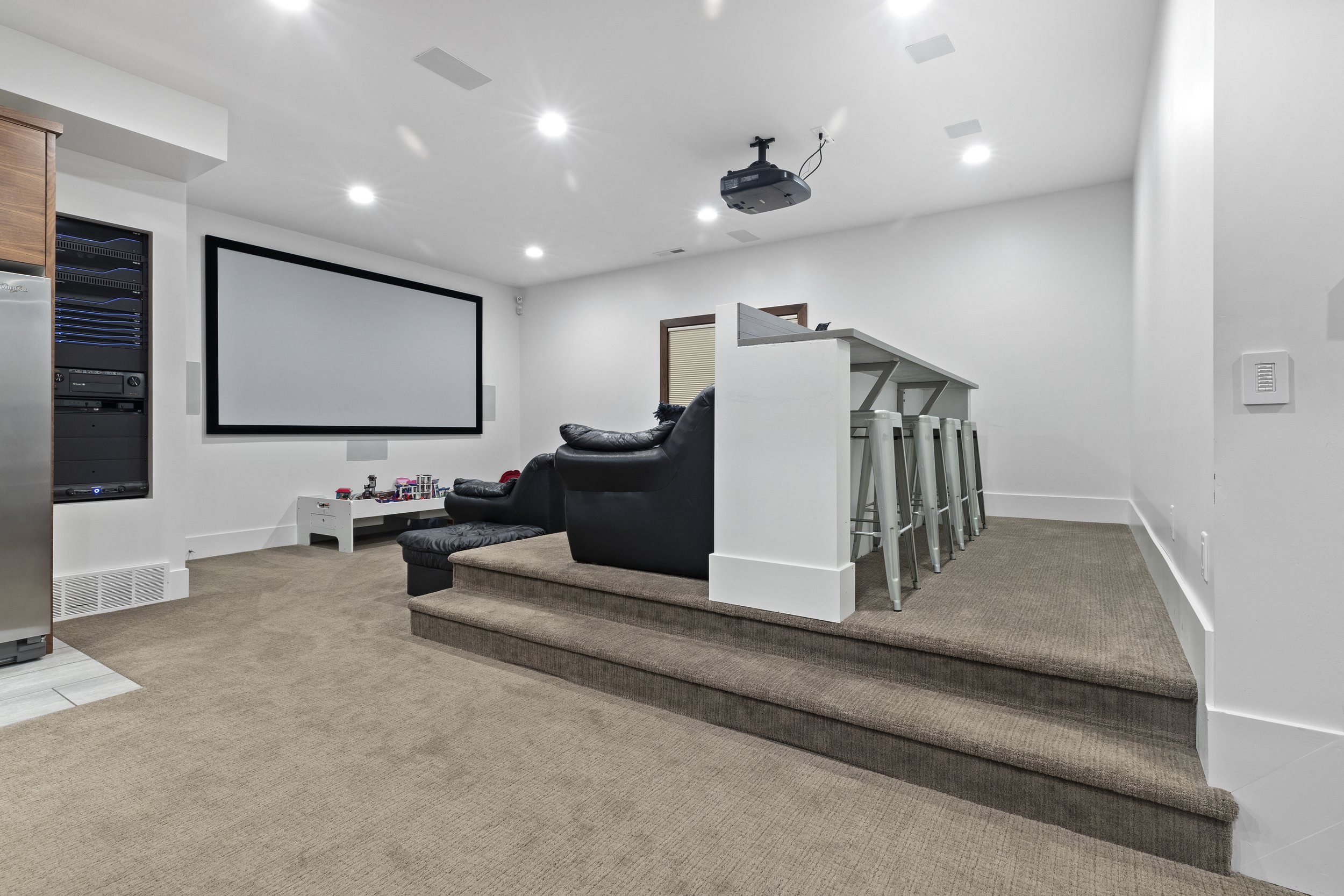 A modern living room with a large white projection screen on the wall, black leather chairs, a small white table with toys, a bar area with barstools, and a ceiling-mounted projector.