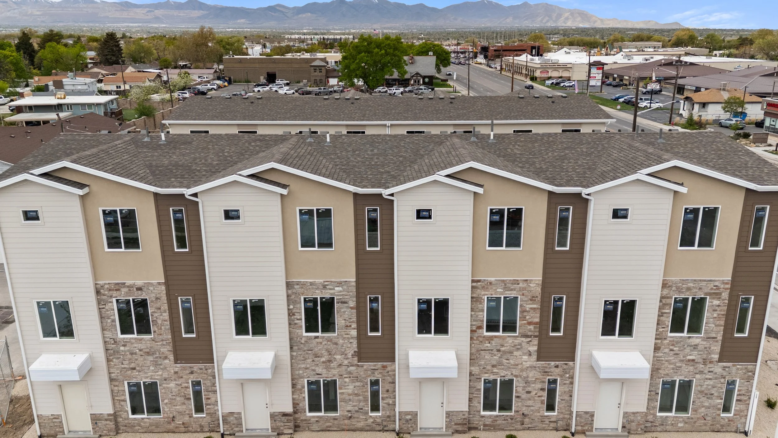 An aerial view of a modern multi-story residential building with a brown and beige exterior, multiple windows, and a stone accent on the lower part of the facade, with a residential neighborhood and commercial area, trees, and mountains in the backgr
