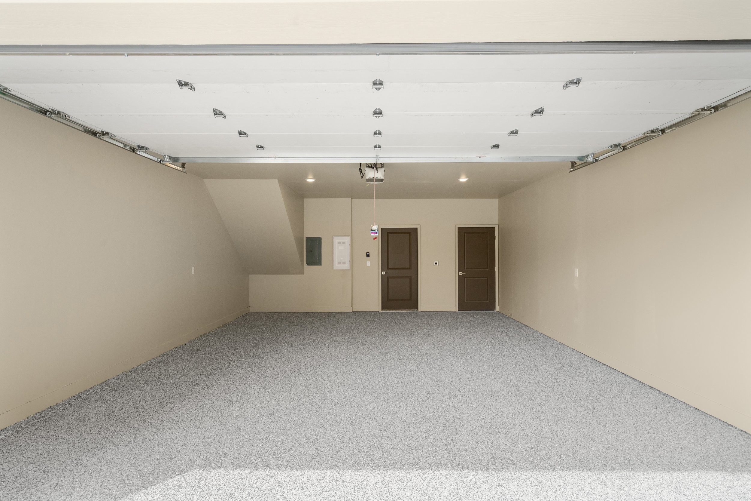 Empty garage with beige walls, gray carpeted floor, brown doors, electrical panel, and garage door openers.