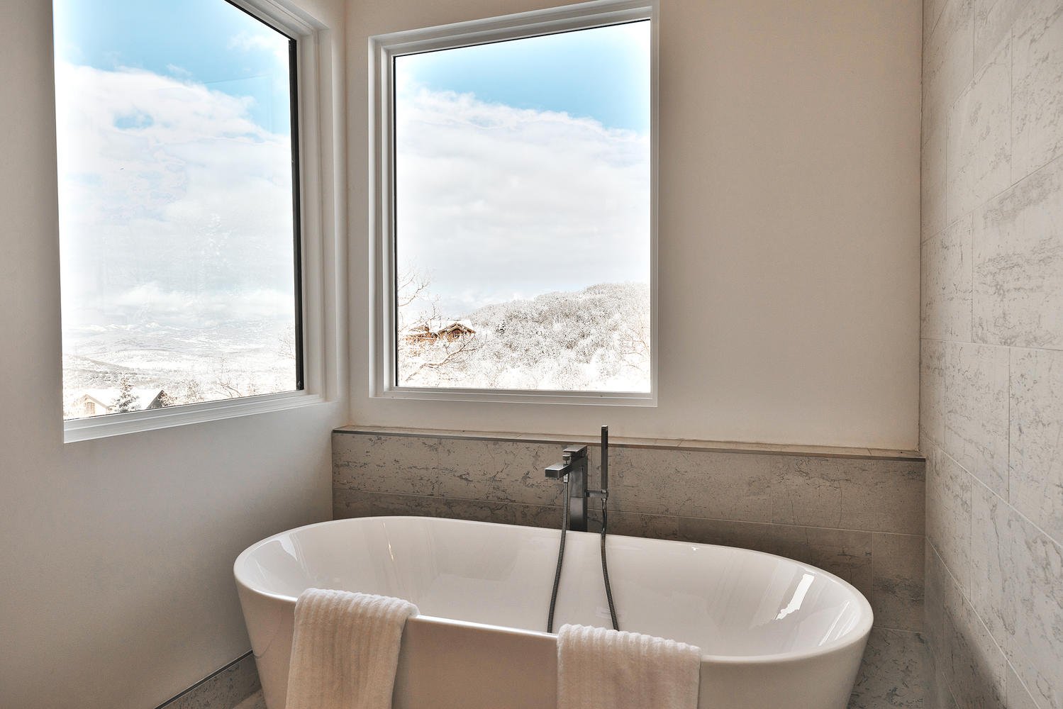 White bathtub with towels draped over the edge, positioned in front of large windows showing a snowy outdoor landscape.