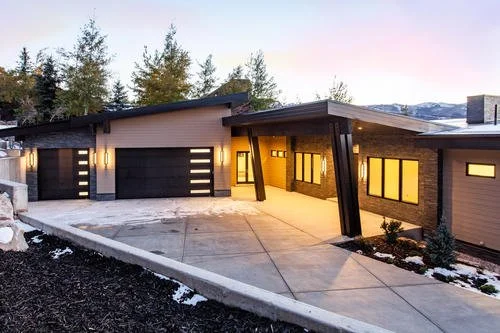 Modern house with a sloped roof, large garage doors, and a driveway, situated in a semi-rural area with trees and snow on the ground.