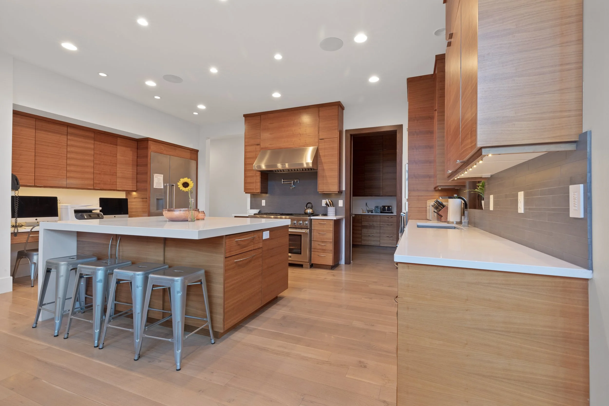 Modern kitchen with wooden cabinets, white countertops, stainless steel appliances, and a kitchen island with three silver bar stools. There is a sunflower in a glass vase on the island and a small wooden knife block on the counter.