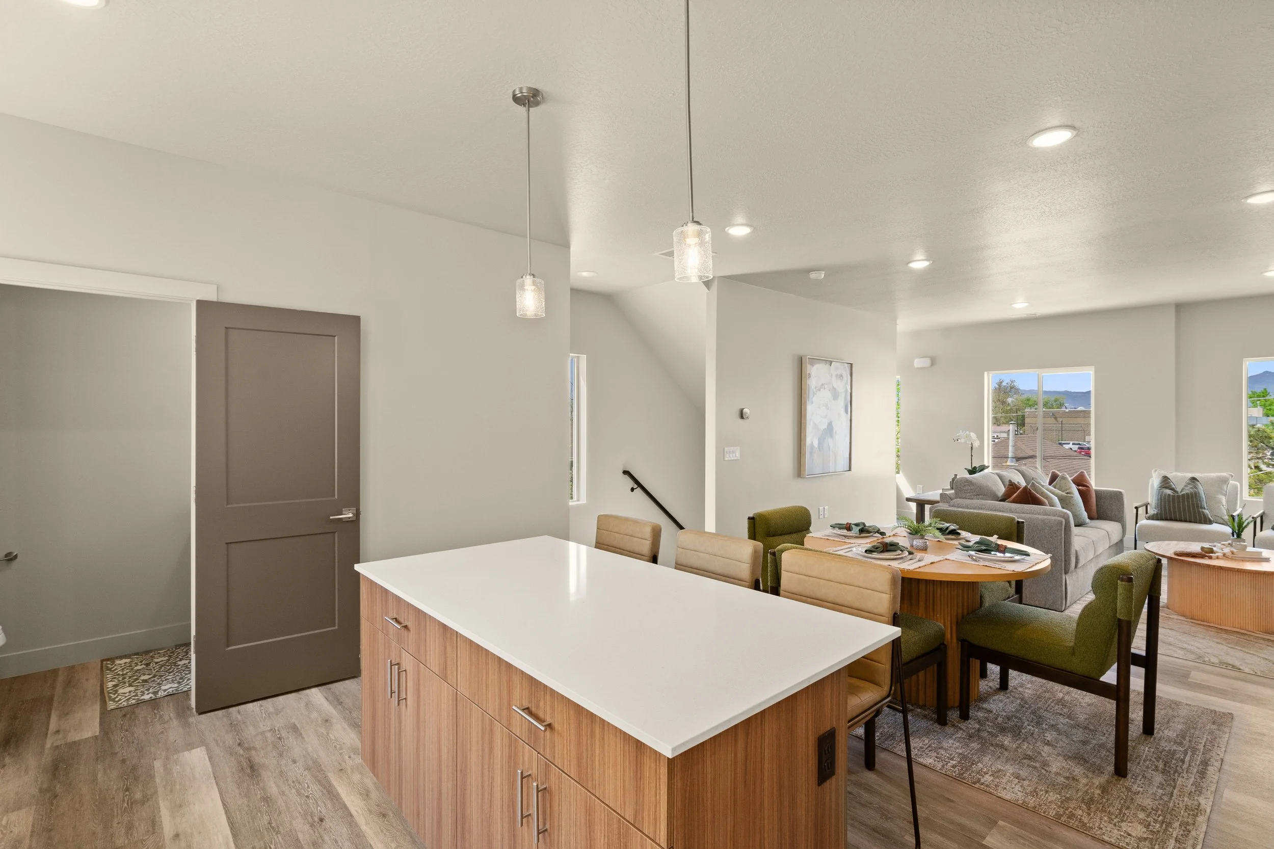 The image shows an open-concept living room and kitchen with a white quartz kitchen island, dining table with beige chairs, and a cozy living area with gray couches and large windows letting in natural light.