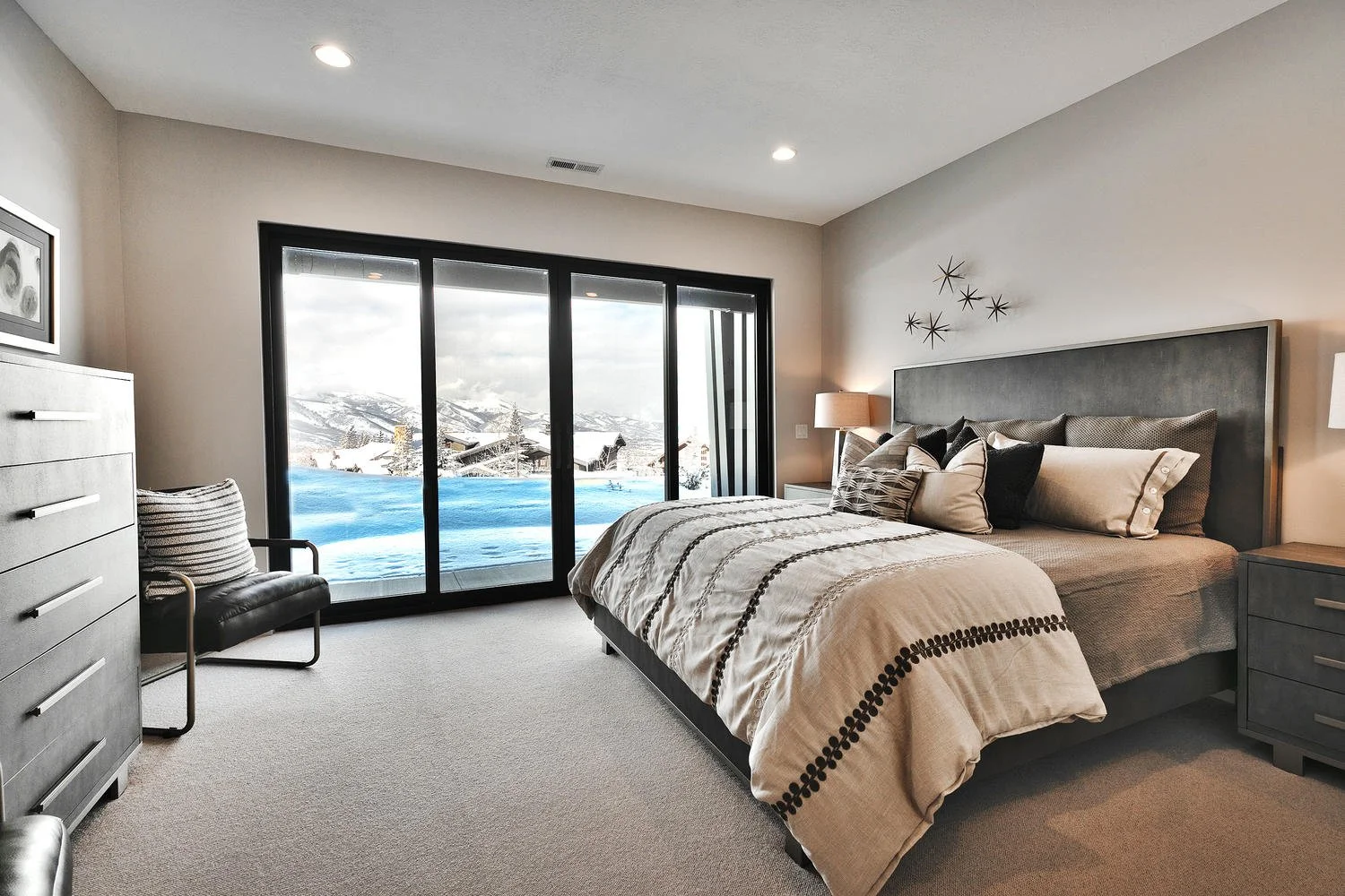 A bedroom with a large window showing a snowy landscape outside, equipped with a bed, nightstand, armchair, and dresser.