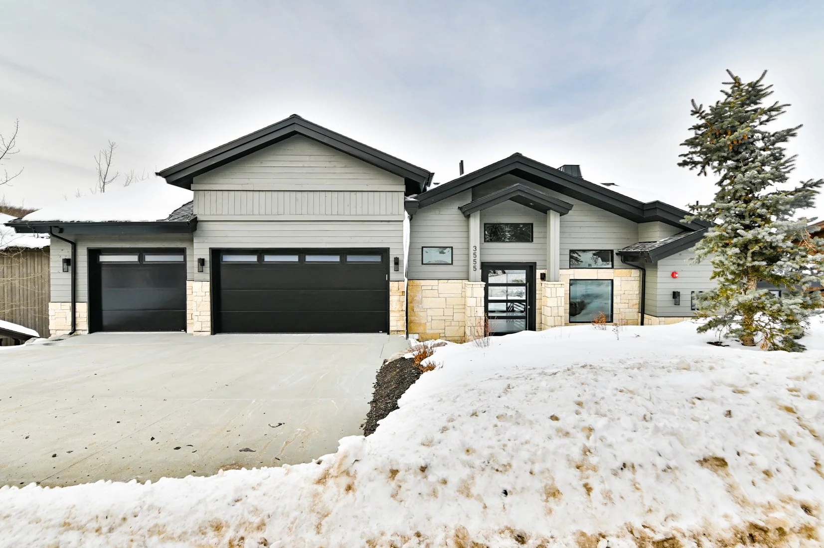 Modern house with gray siding, stone accents, and black garage doors, surrounded by snow and a pine tree to the right.