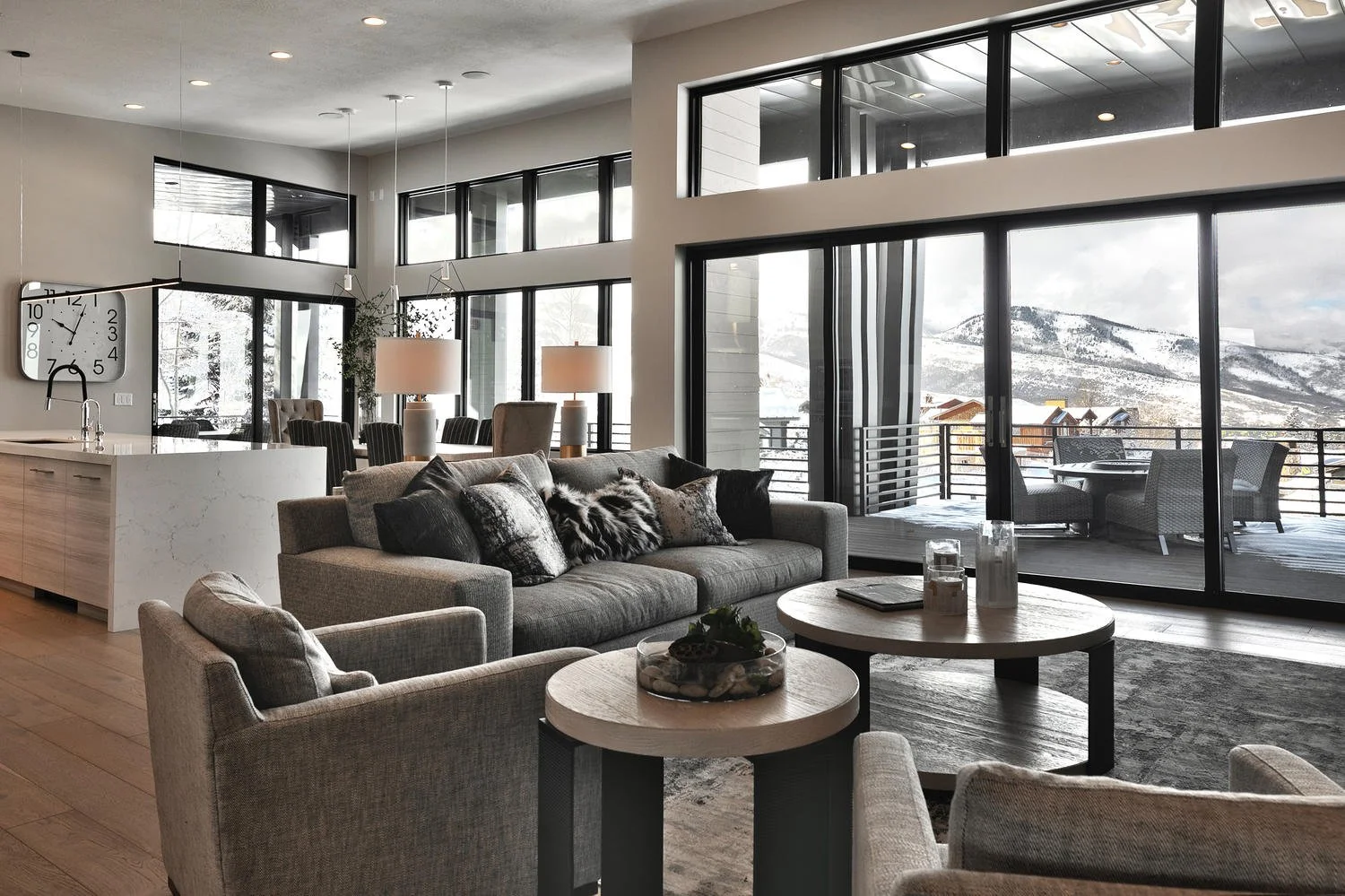 Modern living room with large windows overlooking snowy mountains, gray sofas and armchairs, wooden coffee tables, and a balcony with outdoor seating.