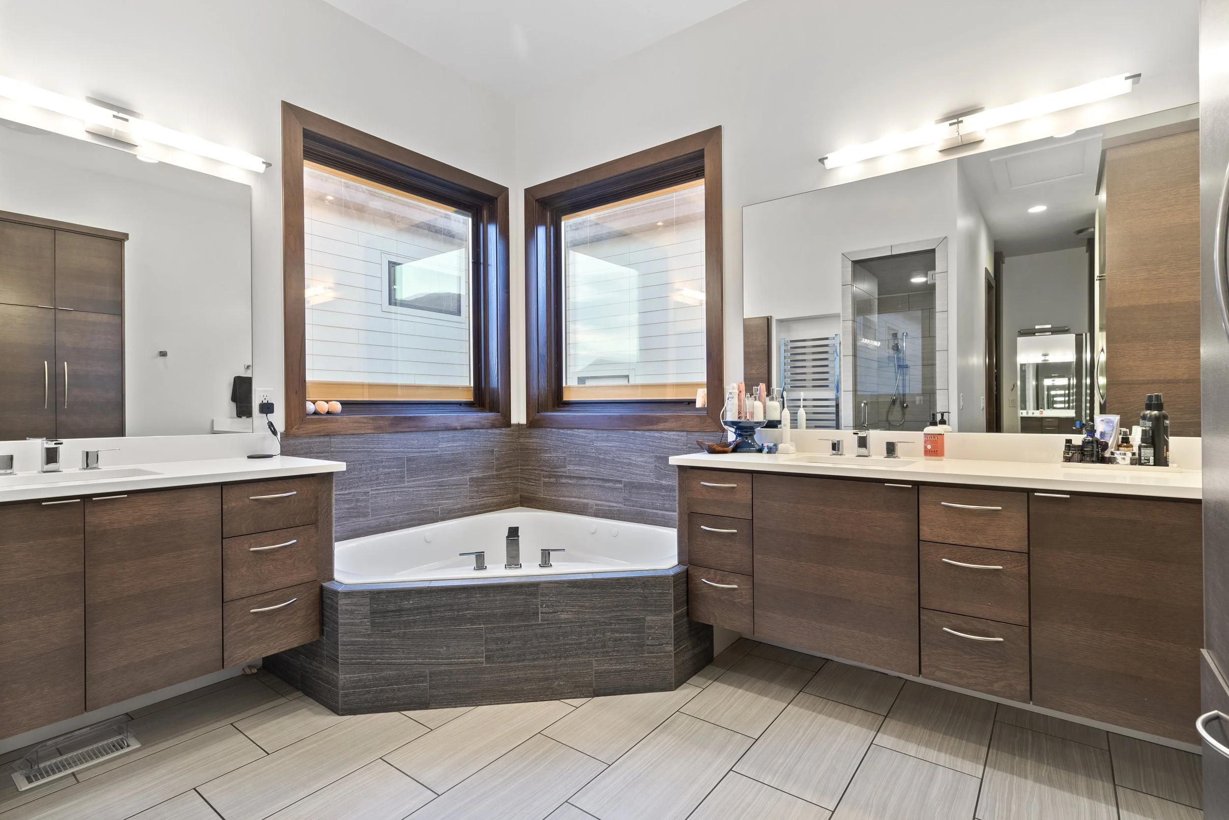 Modern bathroom featuring two sinks with wooden vanities, a large bathtub under two windows, and a walk-in shower with glass doors.