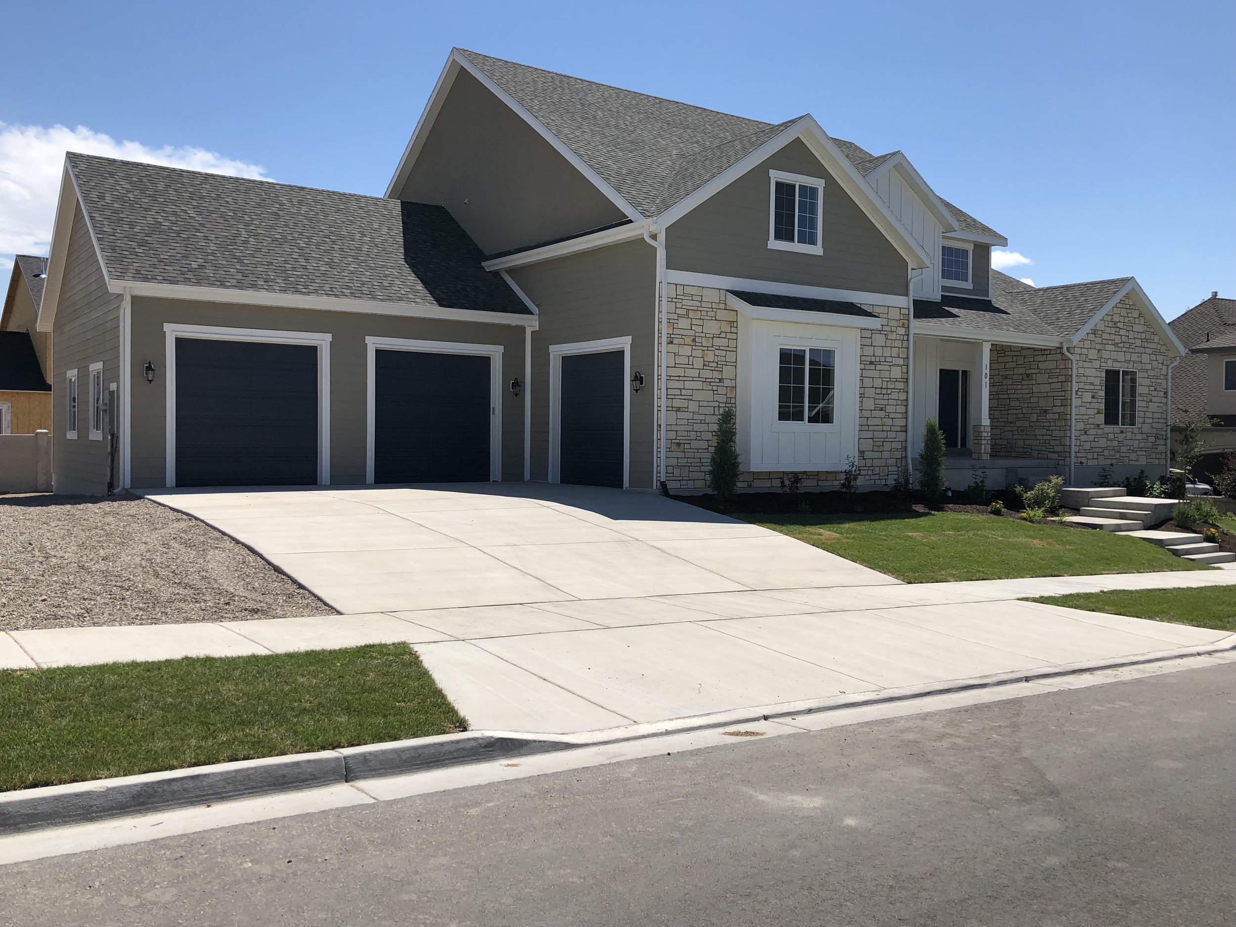 Photo of a modern house with a green and gray exterior, featuring a sloped roof, garage, and front yard with steps and grass under a clear blue sky.