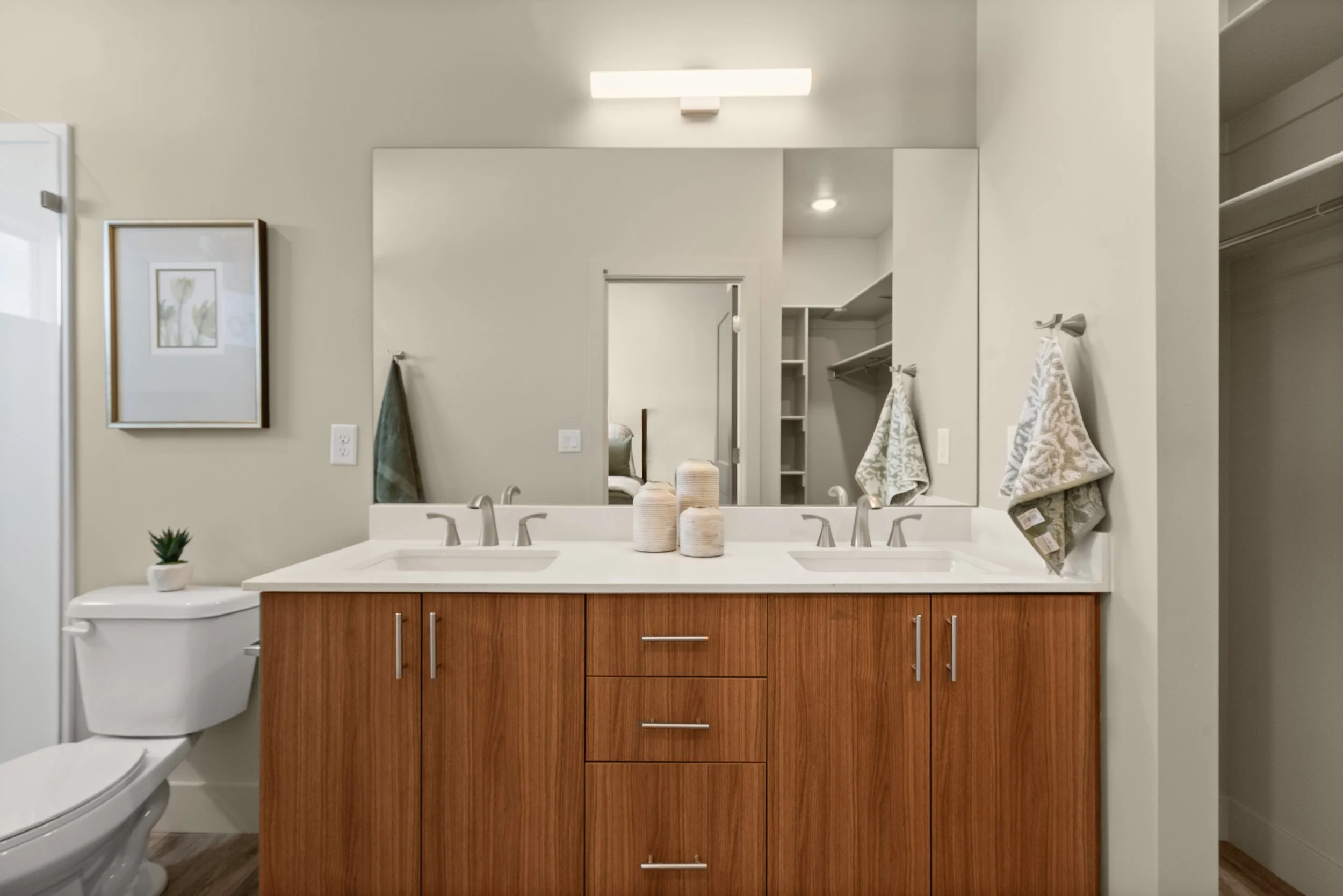 A modern bathroom with a double sink vanity, a large mirror, a framed floral picture, and towels hanging on hooks.